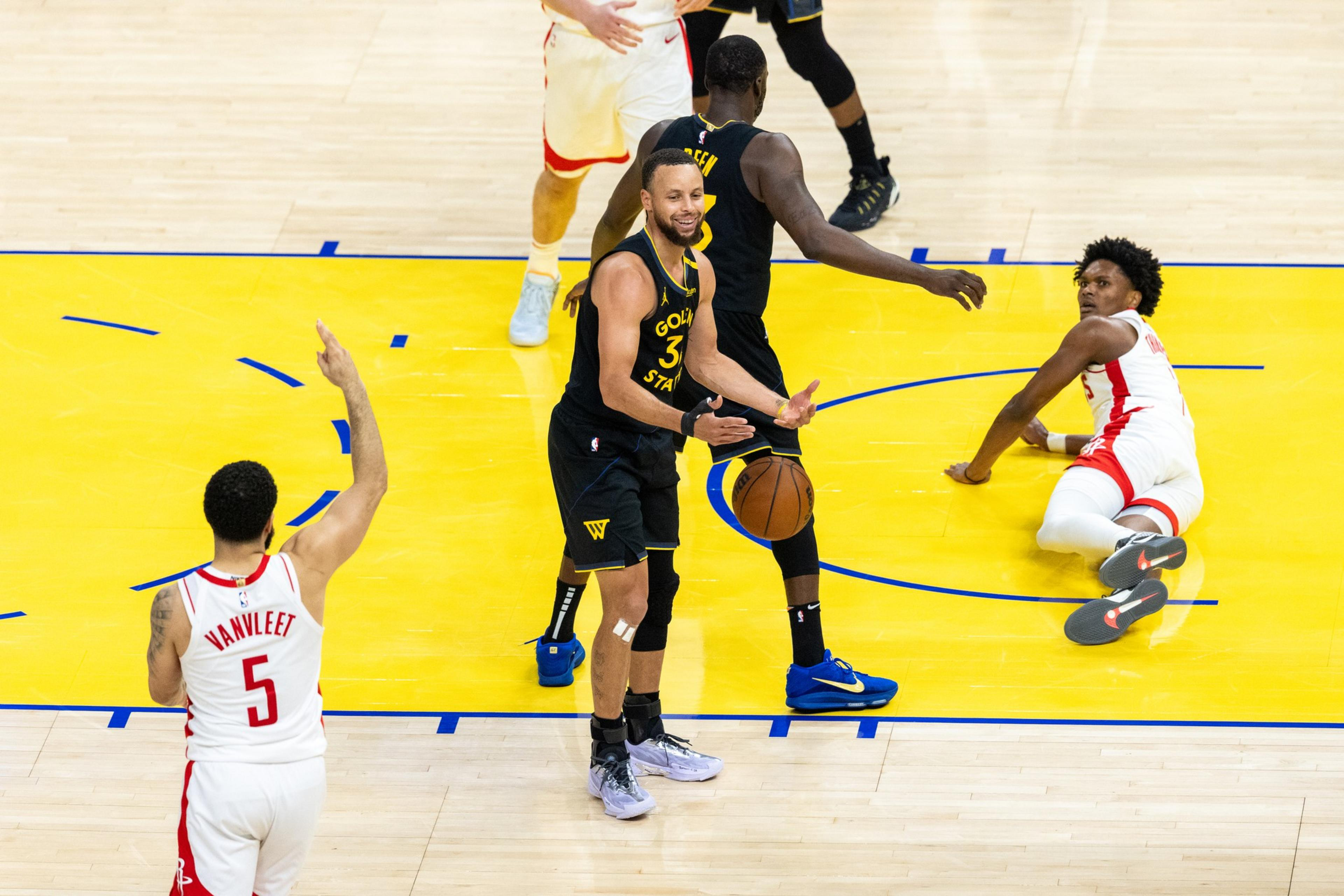 A basketball player in a black uniform is smiling and holding a ball, surrounded by players in red and black jerseys on a brightly lit court.