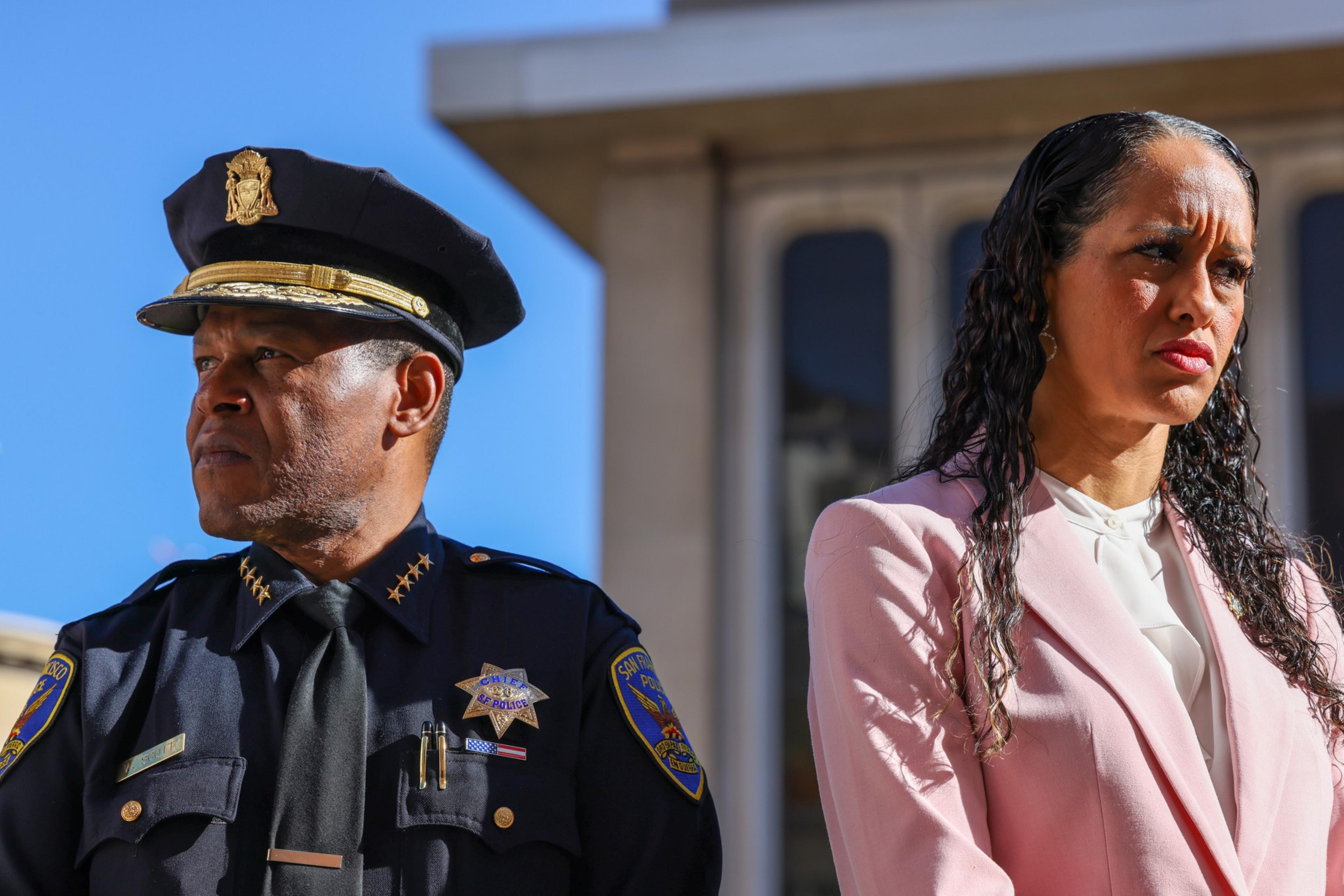 A man in a police uniform and a woman in a pink blazer stand side by side, both looking serious and focused in opposite directions.