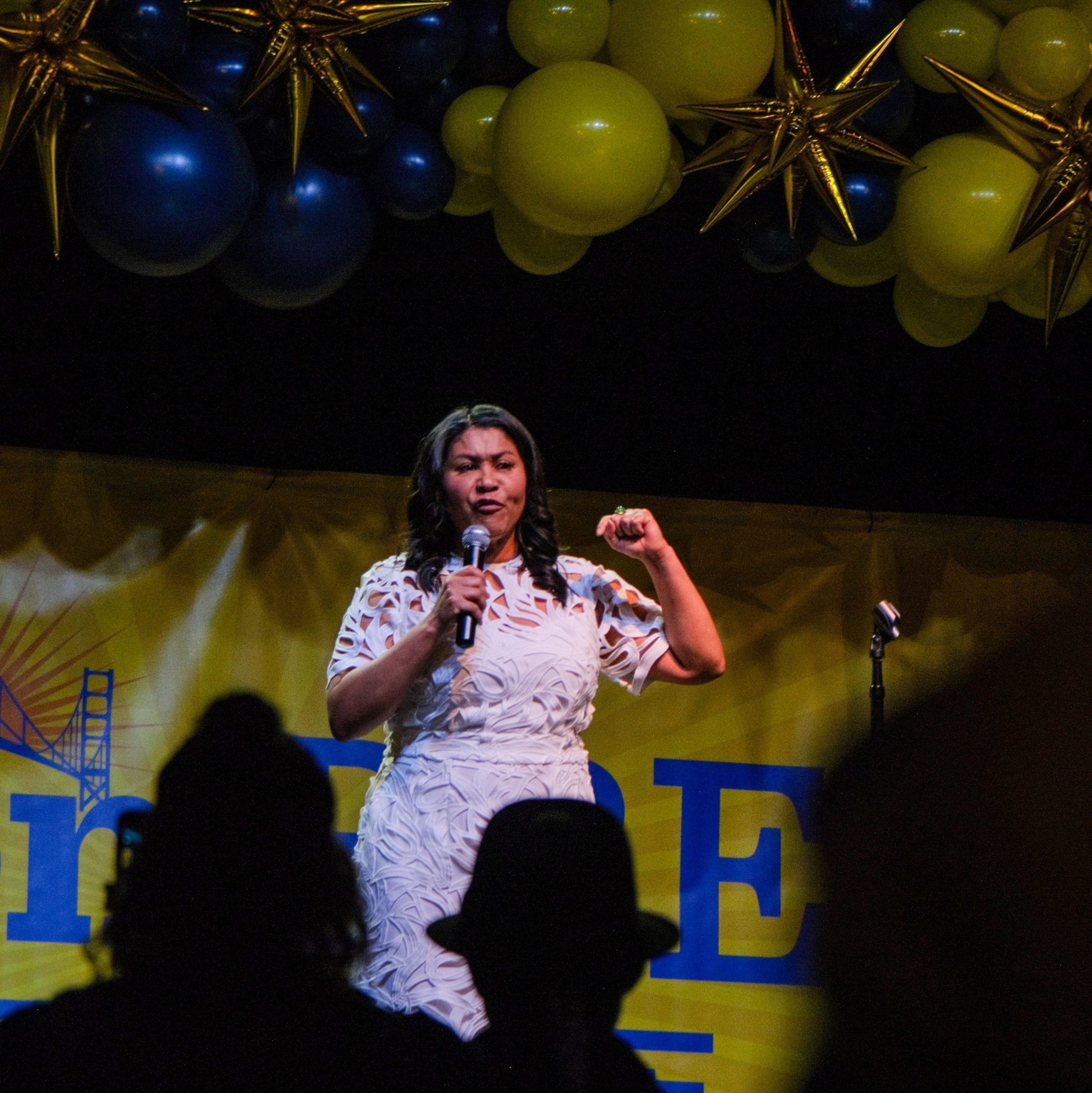 A woman stands on stage holding a microphone, surrounded by yellow and blue balloons. The backdrop shows the Golden Gate Bridge graphic.