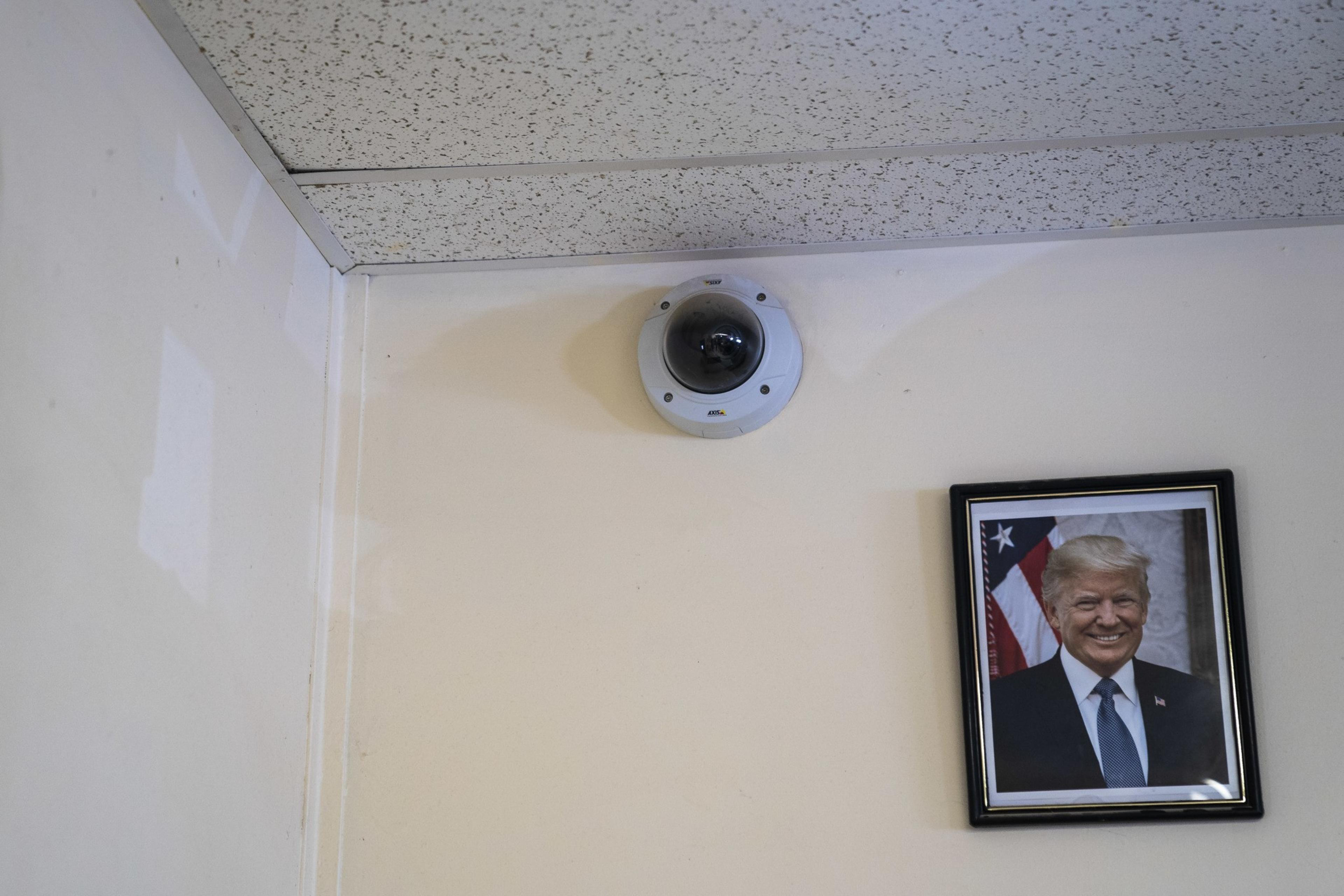A security camera is mounted on a ceiling corner above a framed photo of a man in a suit, smiling, with an American flag visible in the background.