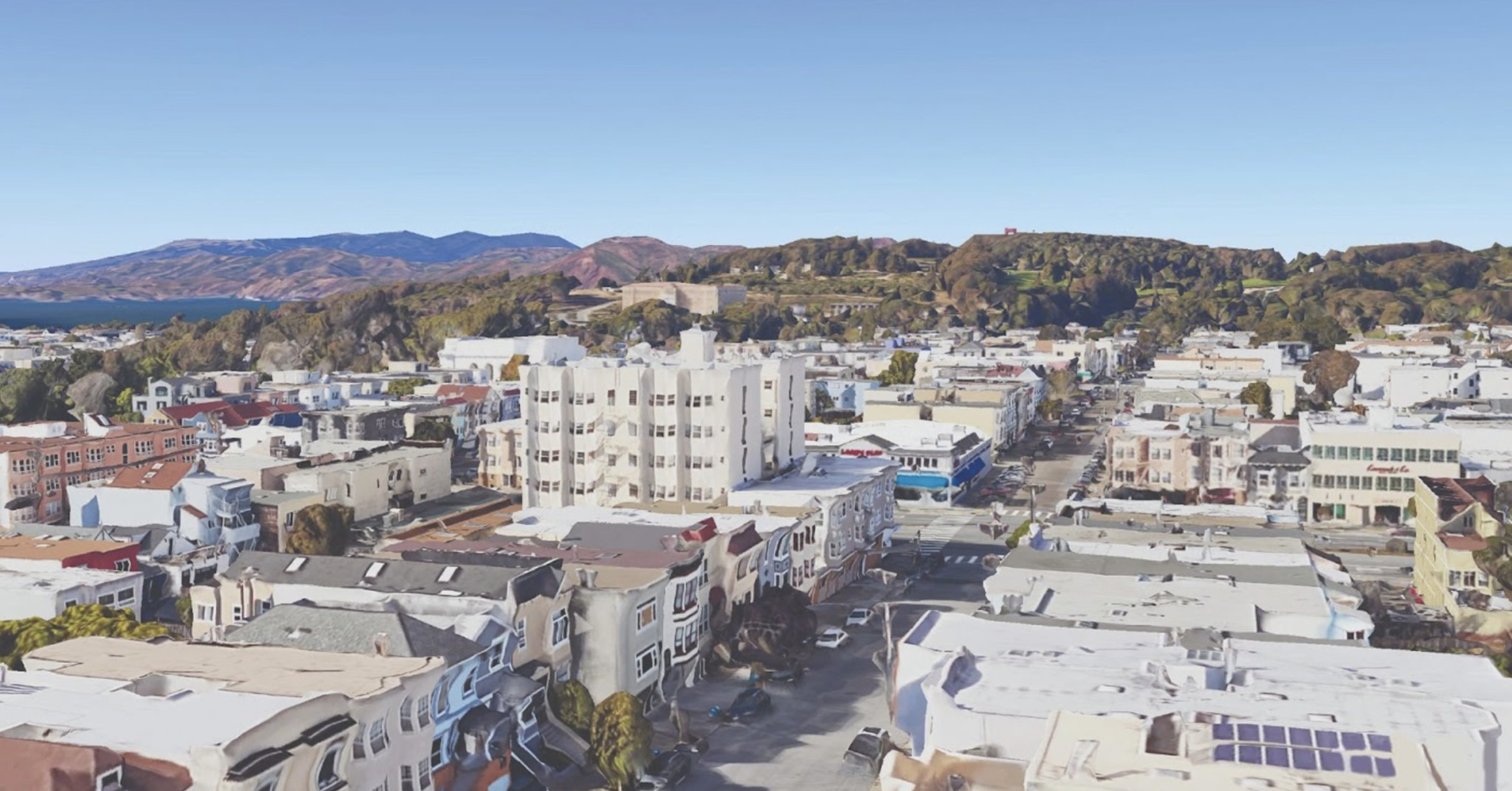 This is an aerial view of a suburban neighborhood with tightly packed buildings, tree-lined streets, and hills in the background under a clear blue sky.