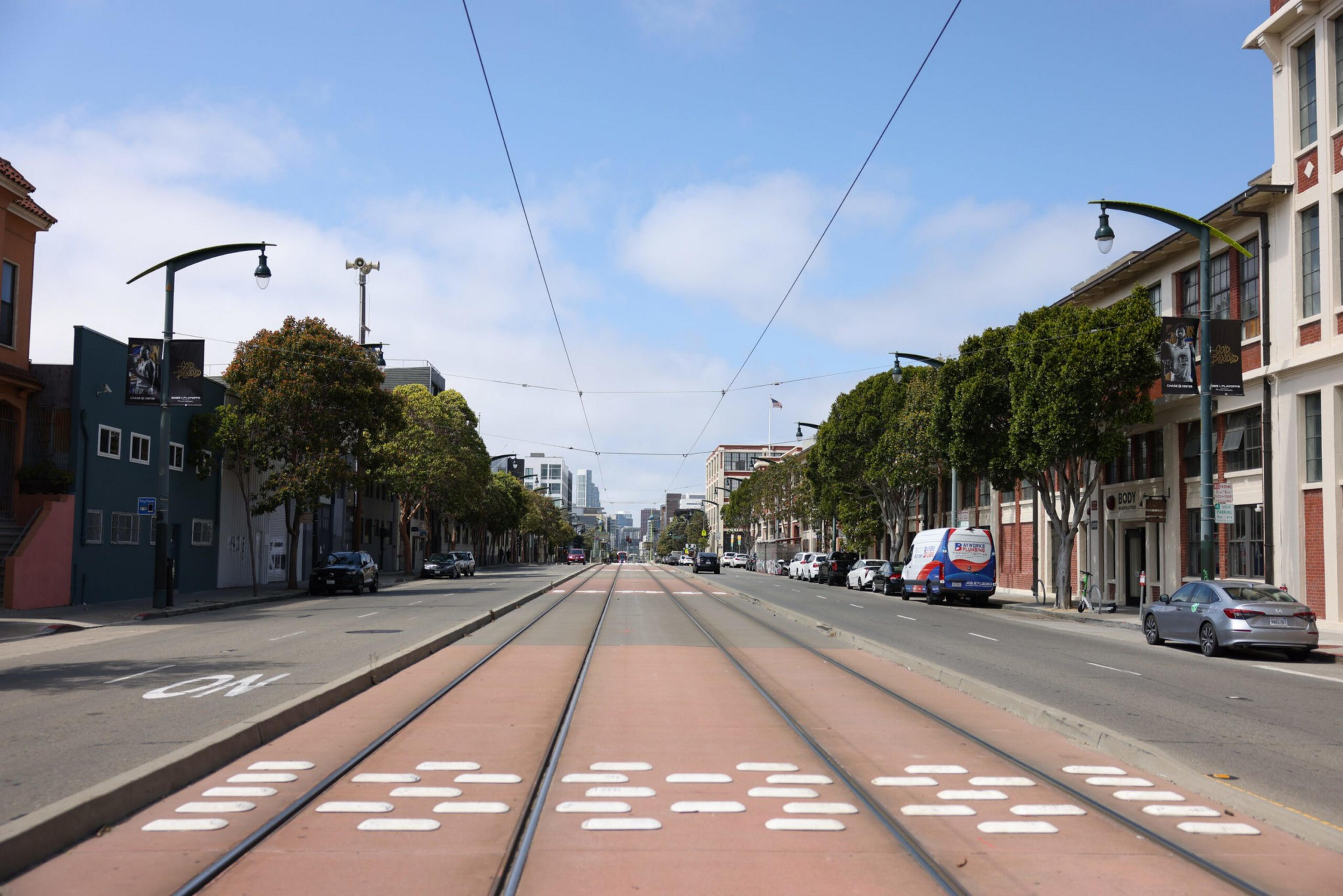 A wide city street features tram tracks in the center, with buildings and trees lining both sides. Few vehicles are parked along the street, and the sky is partly cloudy.