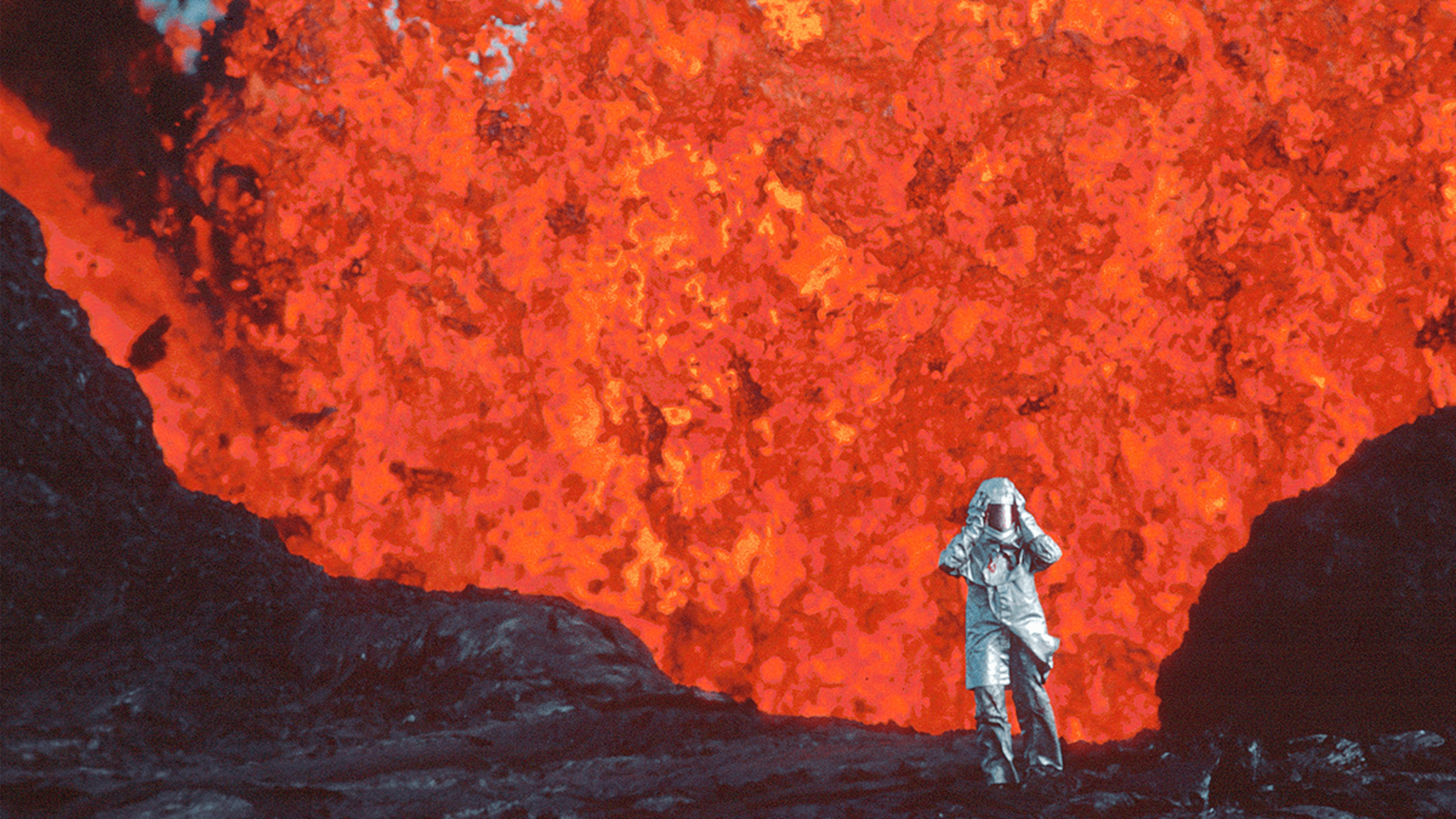 A person in a heat-resistant suit stands near the edge of a glowing, flowing lava field surrounded by dark volcanic rock.