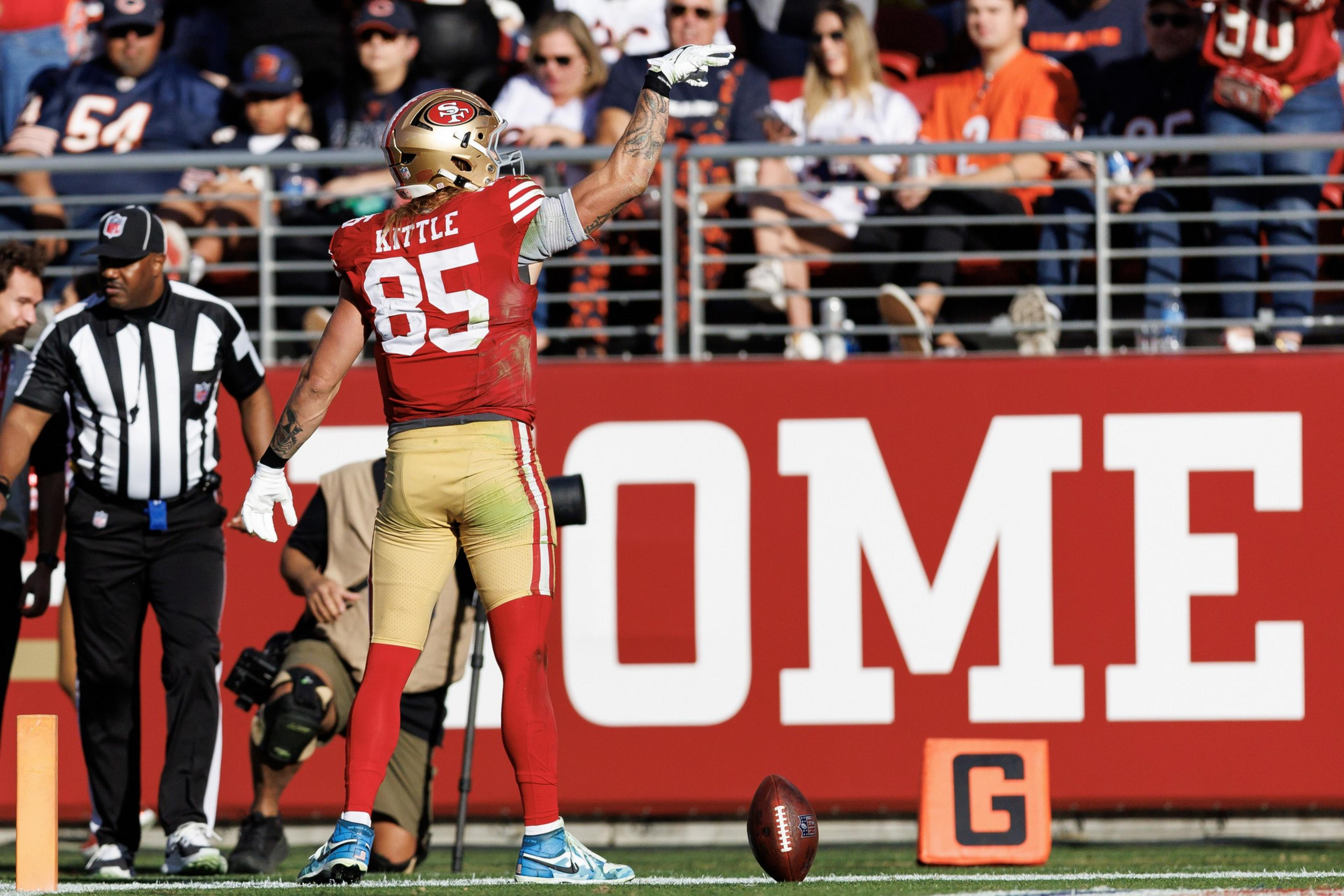 A football player in a red and gold uniform stands on the field gesturing. The number 85 is visible on his jersey. A football is on the ground, with spectators behind.