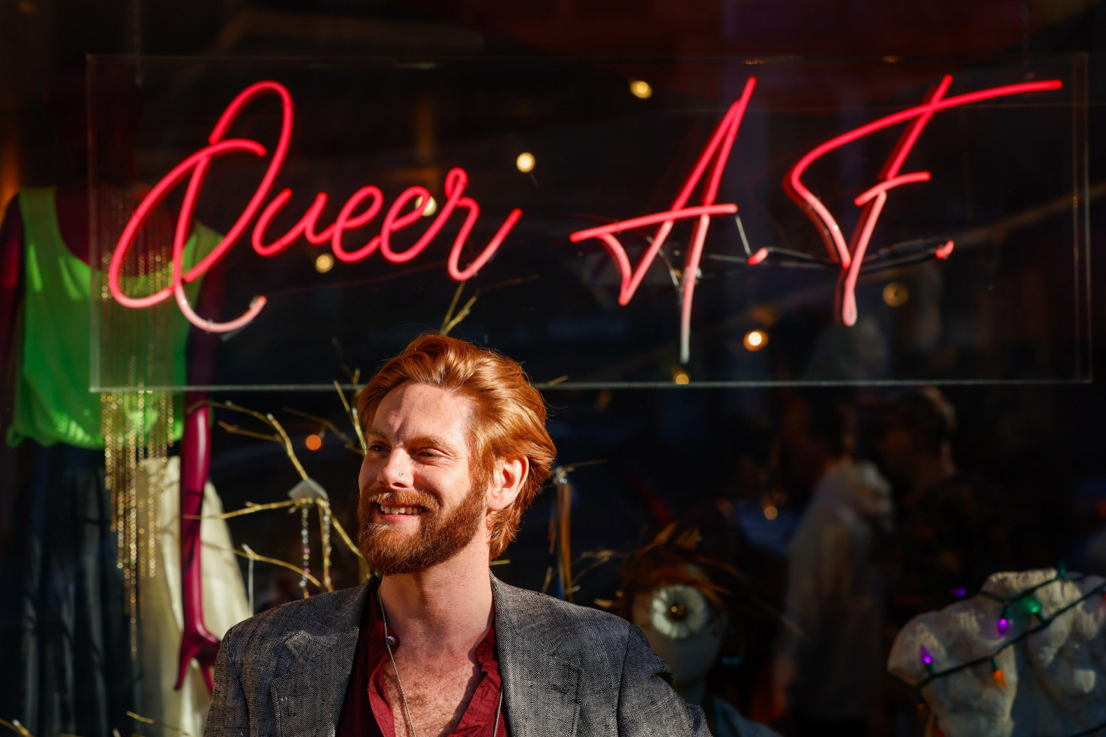 A person with red hair and a beard smiles in front of a neon sign reading "Queer A.F." Bright colors and reflections are visible around them.