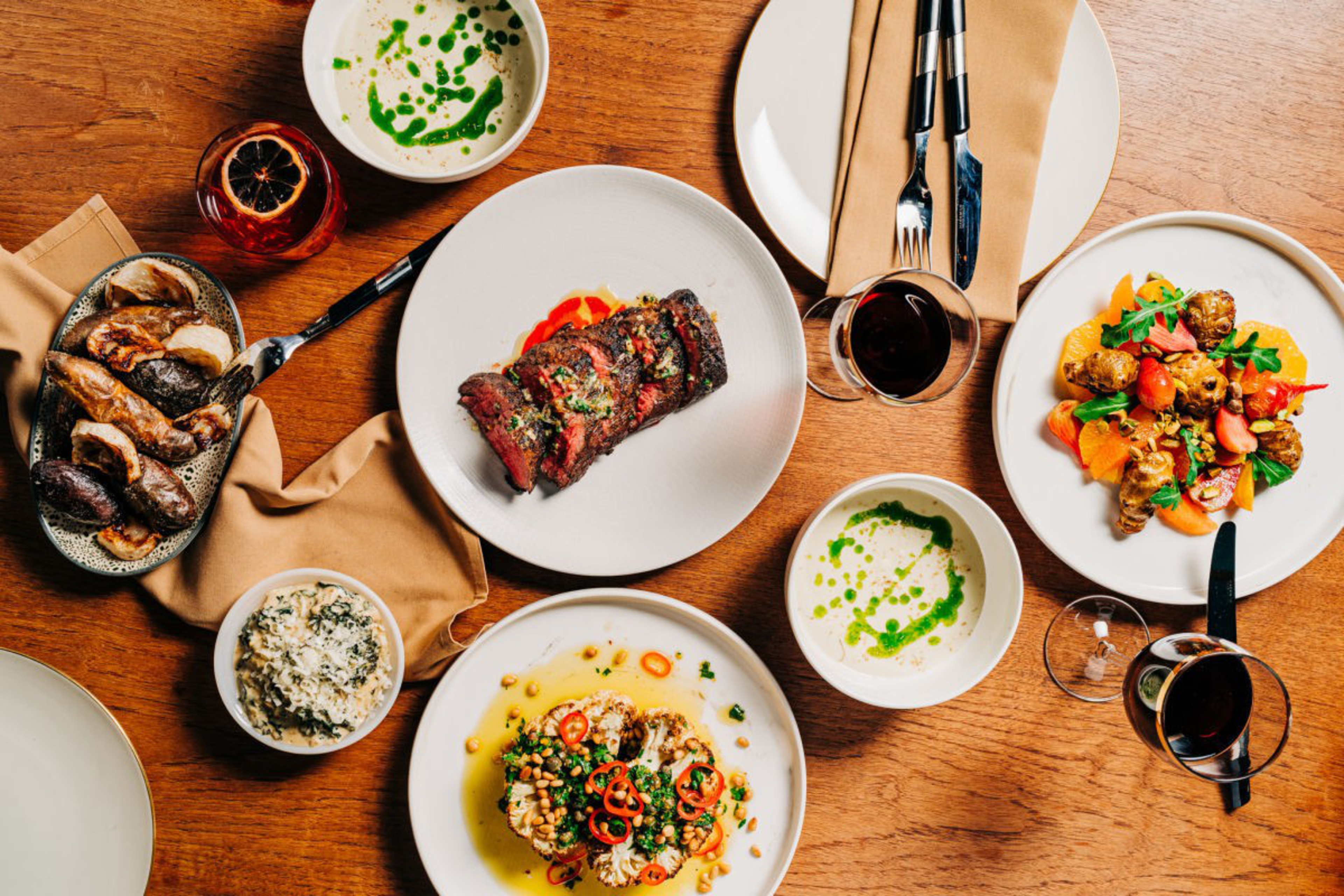 Grilled steak, roasted cauliflower with chili, mixed vegetable salad, mushrooms, creamy soup with herb oil, and two glasses of red wine on a wooden table.
