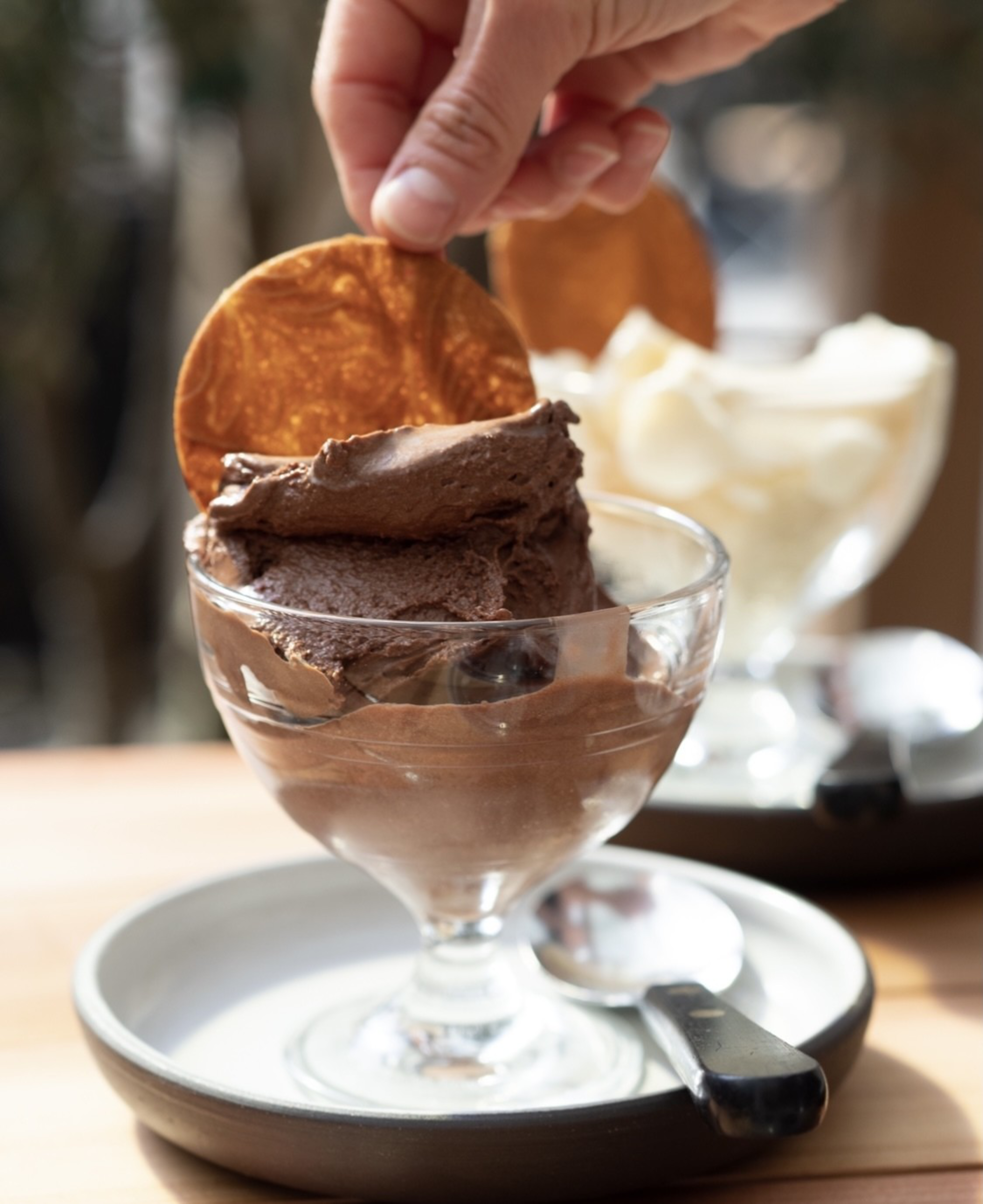 A hand holds a thin, round, golden-brown wafer placed on top of smooth, rich-looking chocolate mousse in a glass dish, with a spoon beside it.