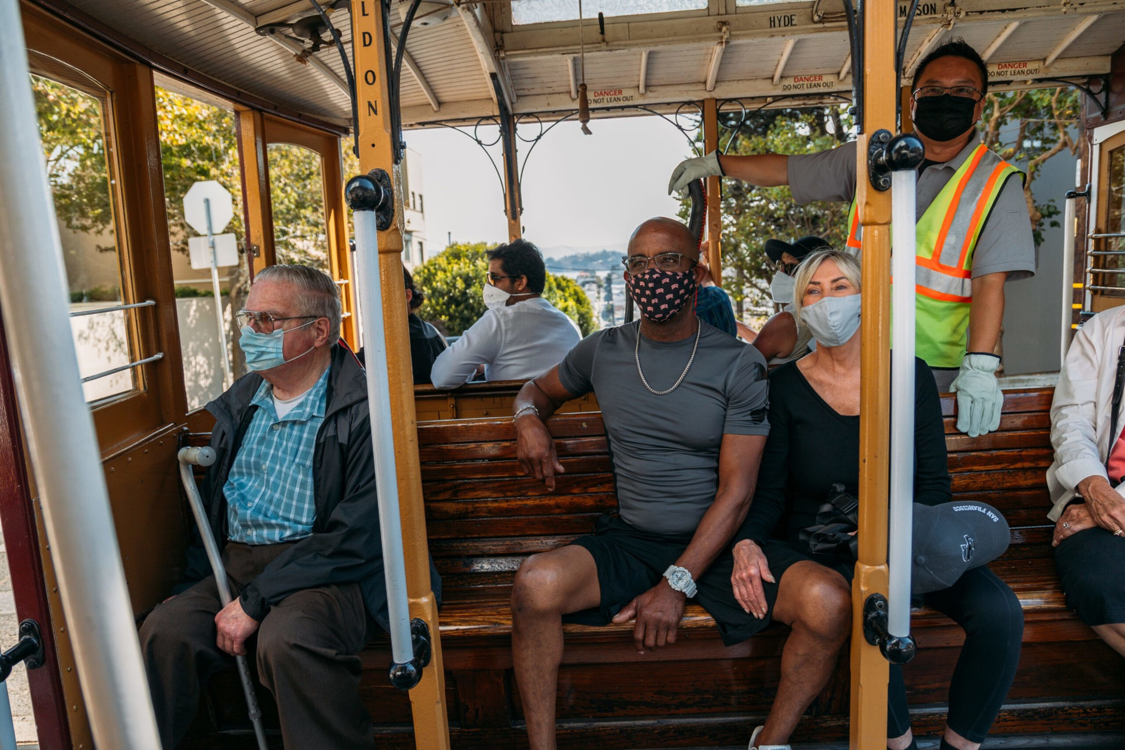 Several people wearing masks sit on wooden benches inside a streetcar, with a standing worker in a safety vest and gloves nearby.
