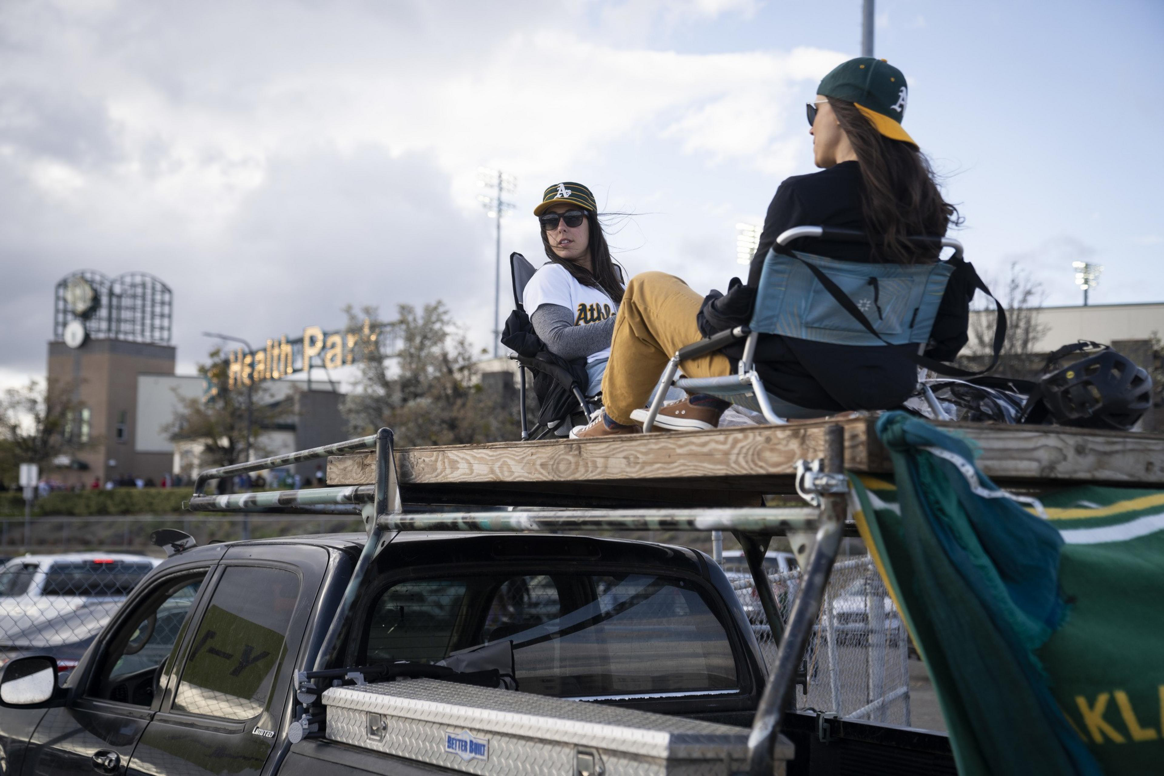 Two people sit in chairs on a truck bed with a makeshift platform, wearing matching sports caps and attire. A stadium and sky are in the background.