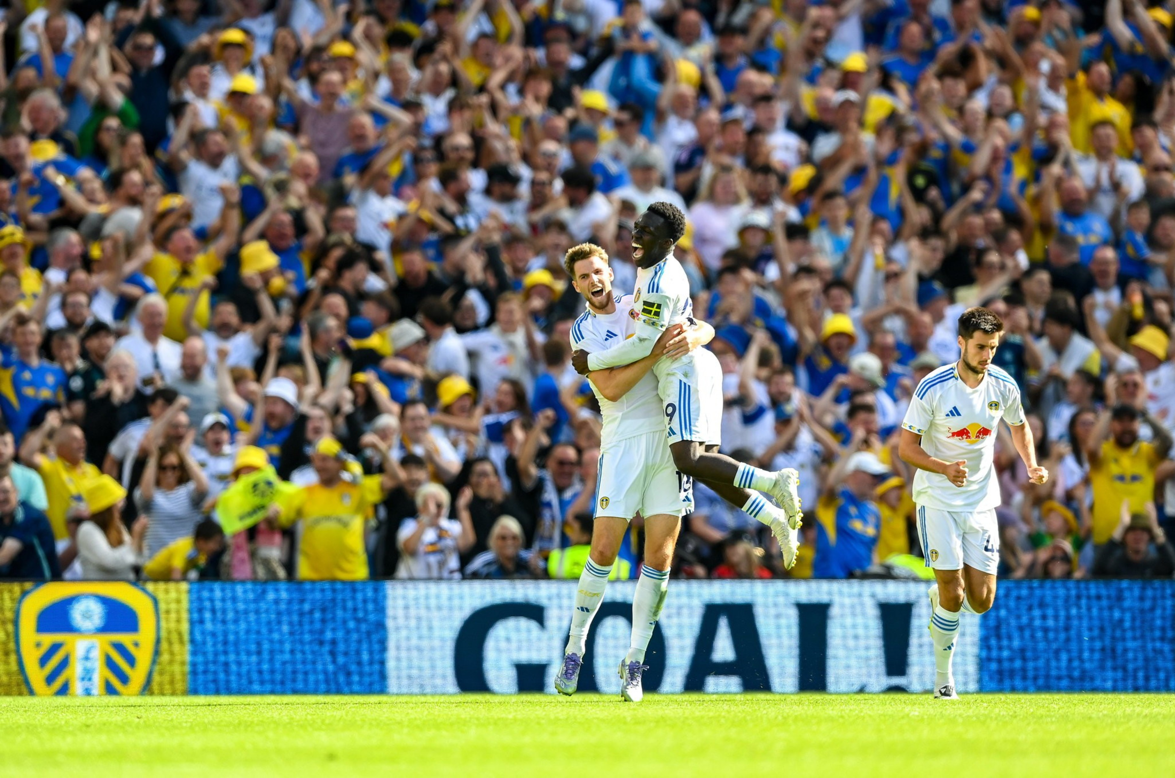 Two soccer players in white jerseys celebrate by hugging as one jumps into the other's arms, with a cheering crowd in yellow and blue in the background.