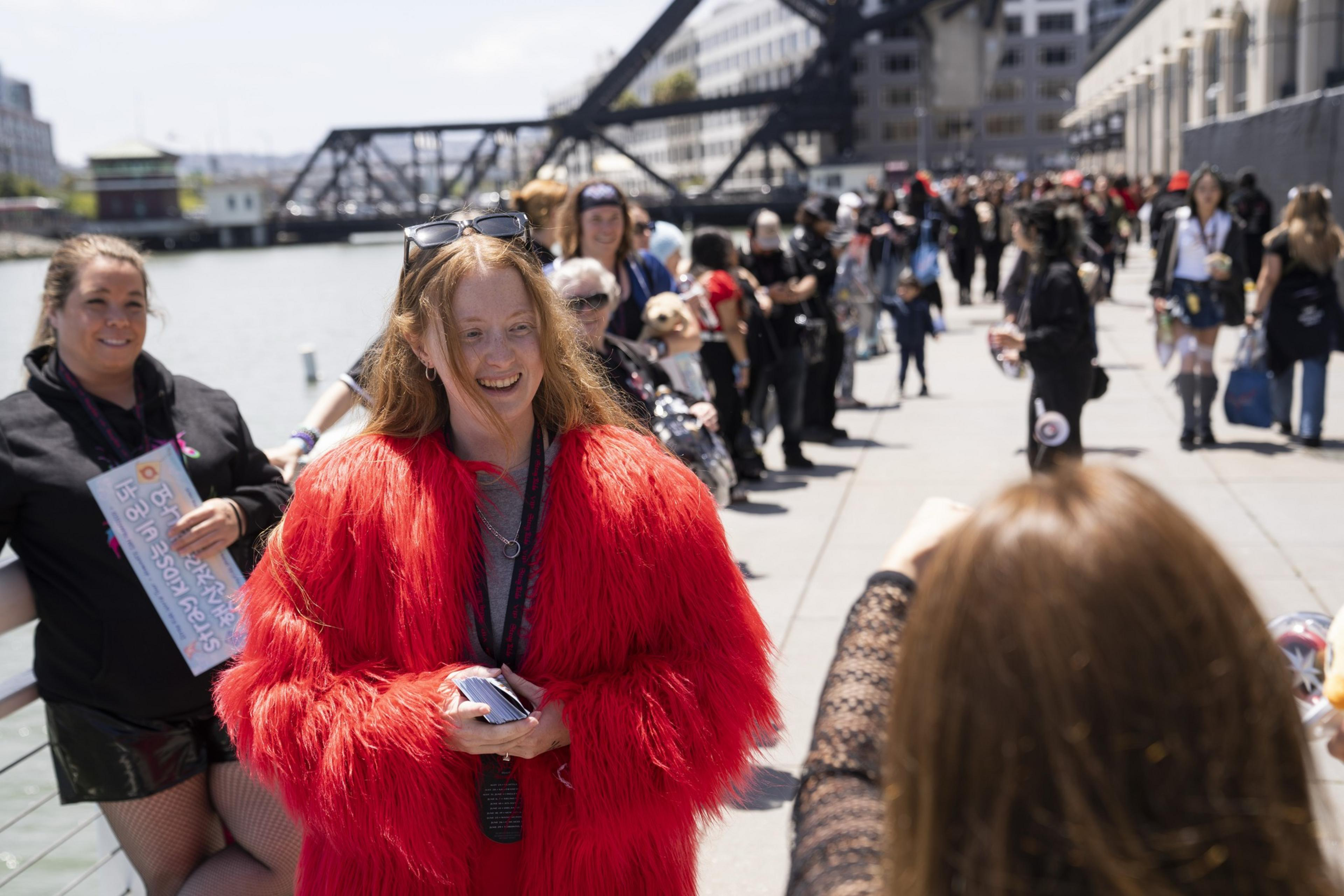 A smiling person in a bright red coat is posing for a photo near water. Behind them, people in various outfits form a long line on a sunny day.