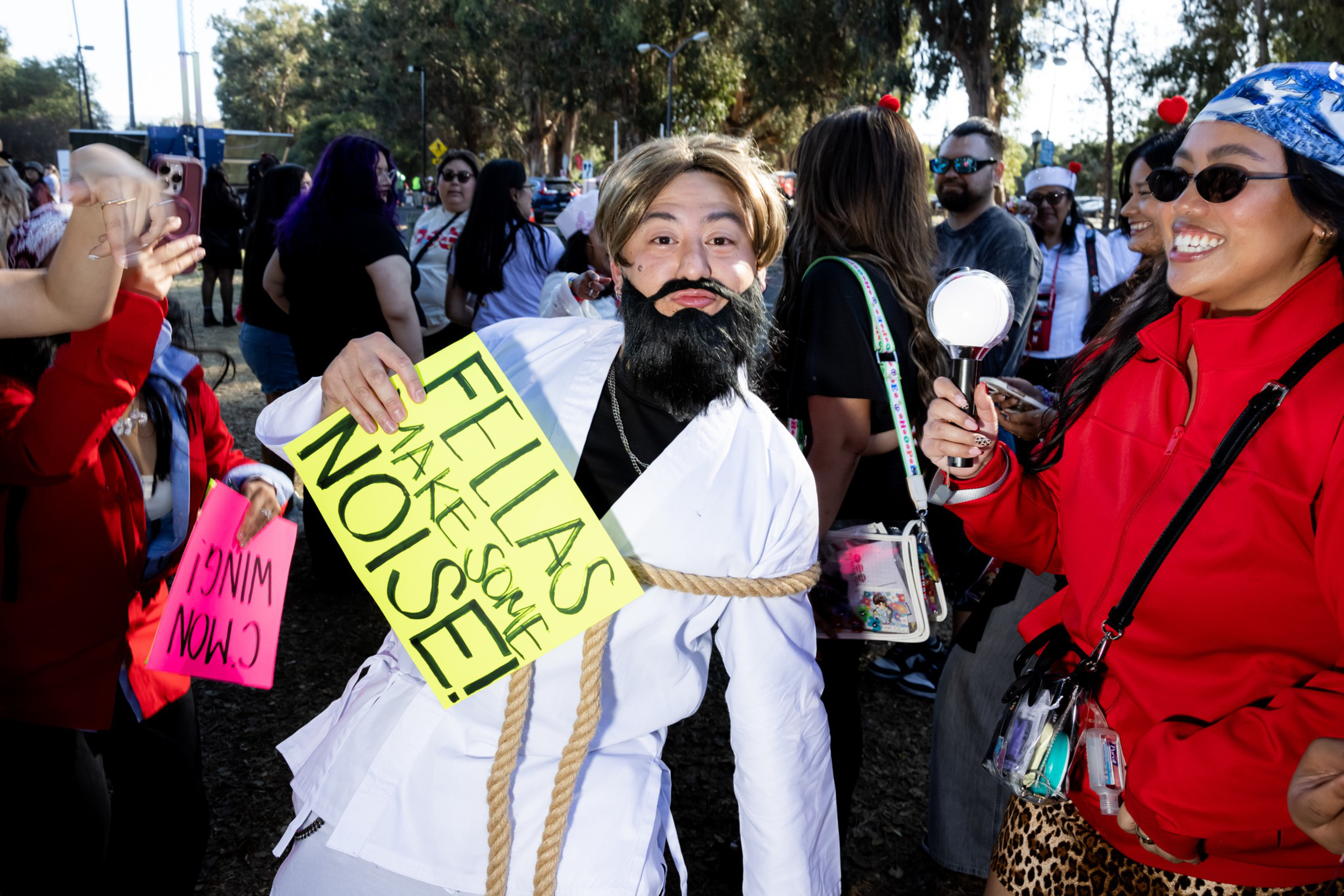 A person dressed as a bearded figure holds a sign saying “FELLAS MAKE SOME NOISE!” while others around them laugh and cheer outdoors.