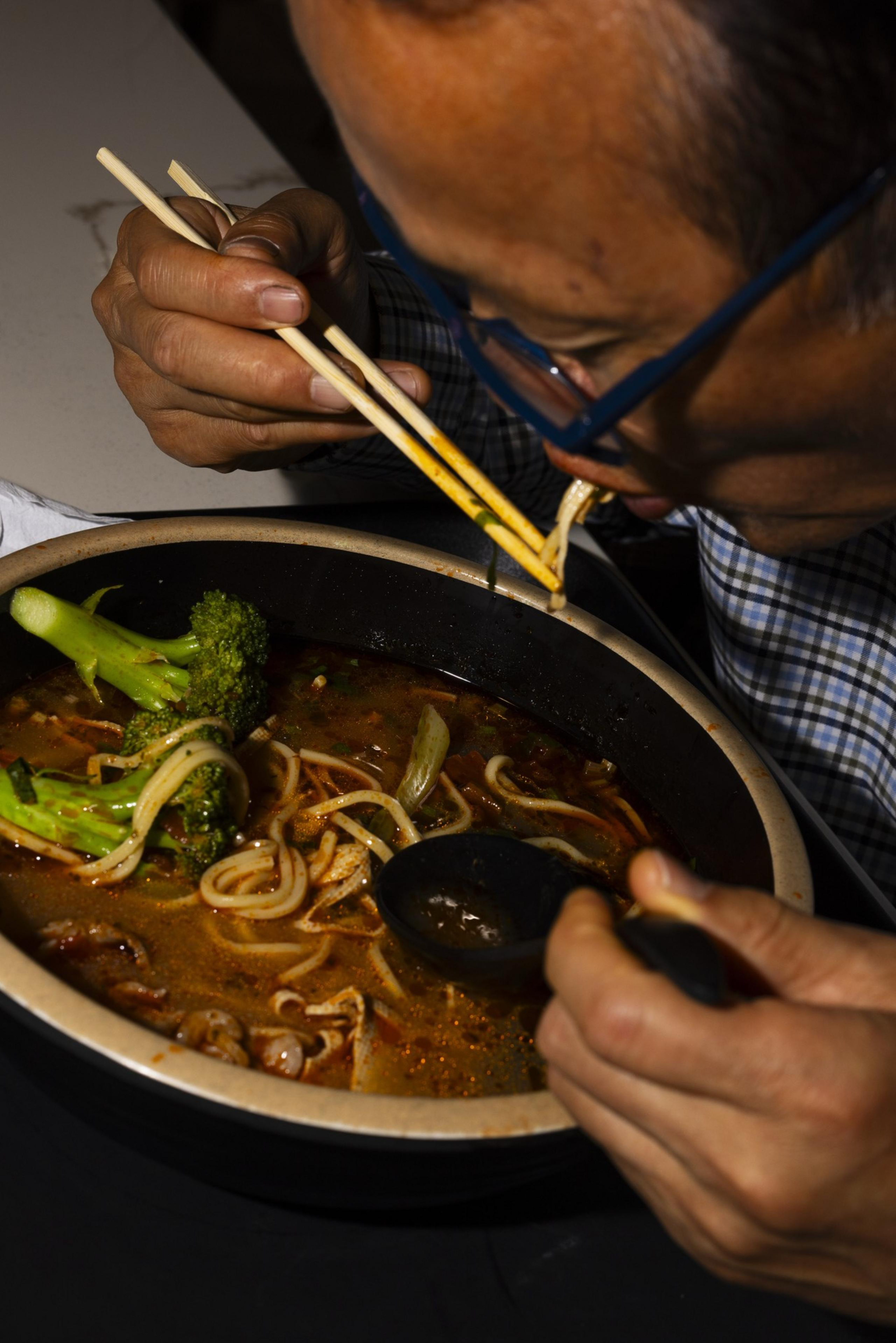 A person is using chopsticks to eat noodles from a bowl filled with broth, noodles, and broccoli, while holding a black spoon in the other hand.