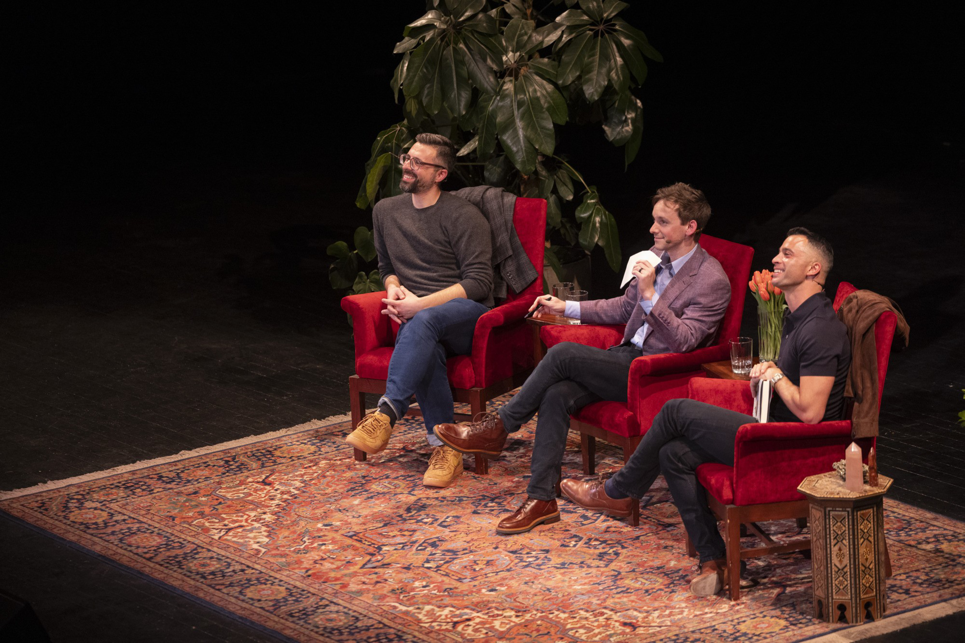 Three people sit on a stage in red armchairs, surrounded by plants and dÃ©cor. They are smiling, engaged in conversation, with a cozy, intimate setting.