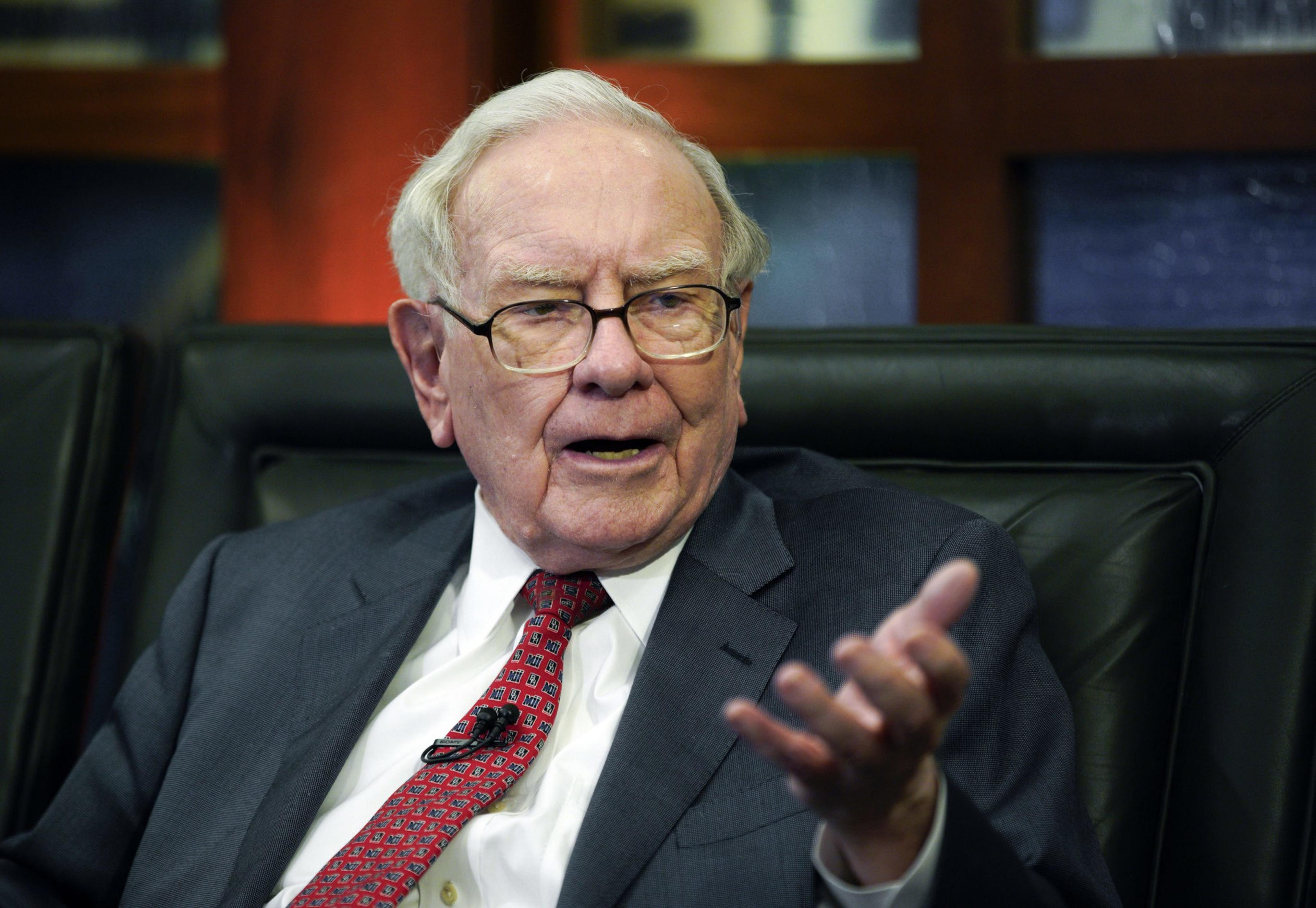An elderly man in a suit speaks animatedly, seated in a leather chair, against a backdrop of bookshelves and framed art.