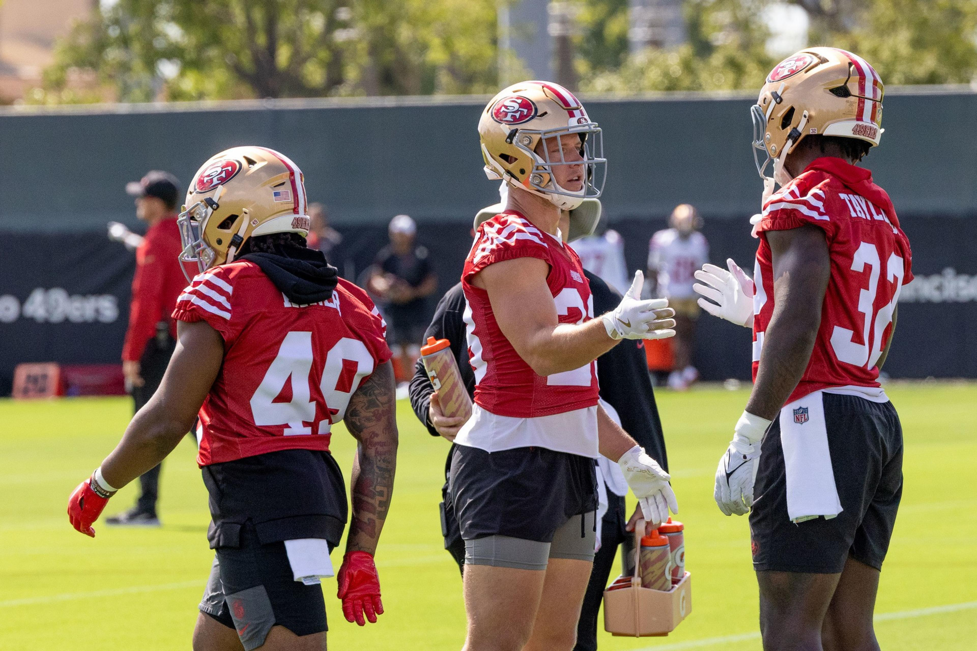 Three football players wear helmets and red jerseys, standing on a field conversing. One holds water bottles, with trees and people in the background.