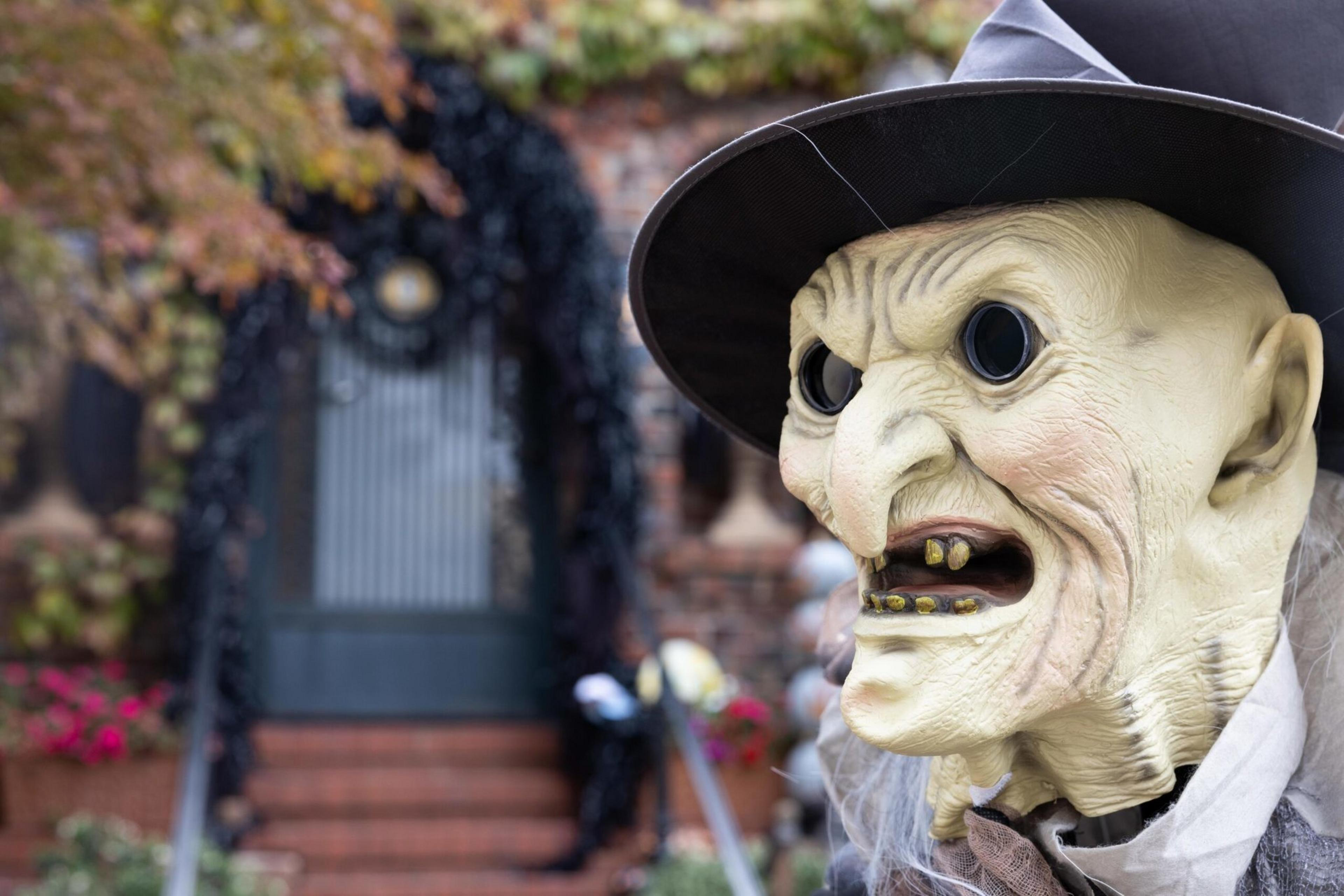 A scary witch mask with hollow black eyes, crooked teeth, and a black pointed hat stands in front of a decorated house entrance.