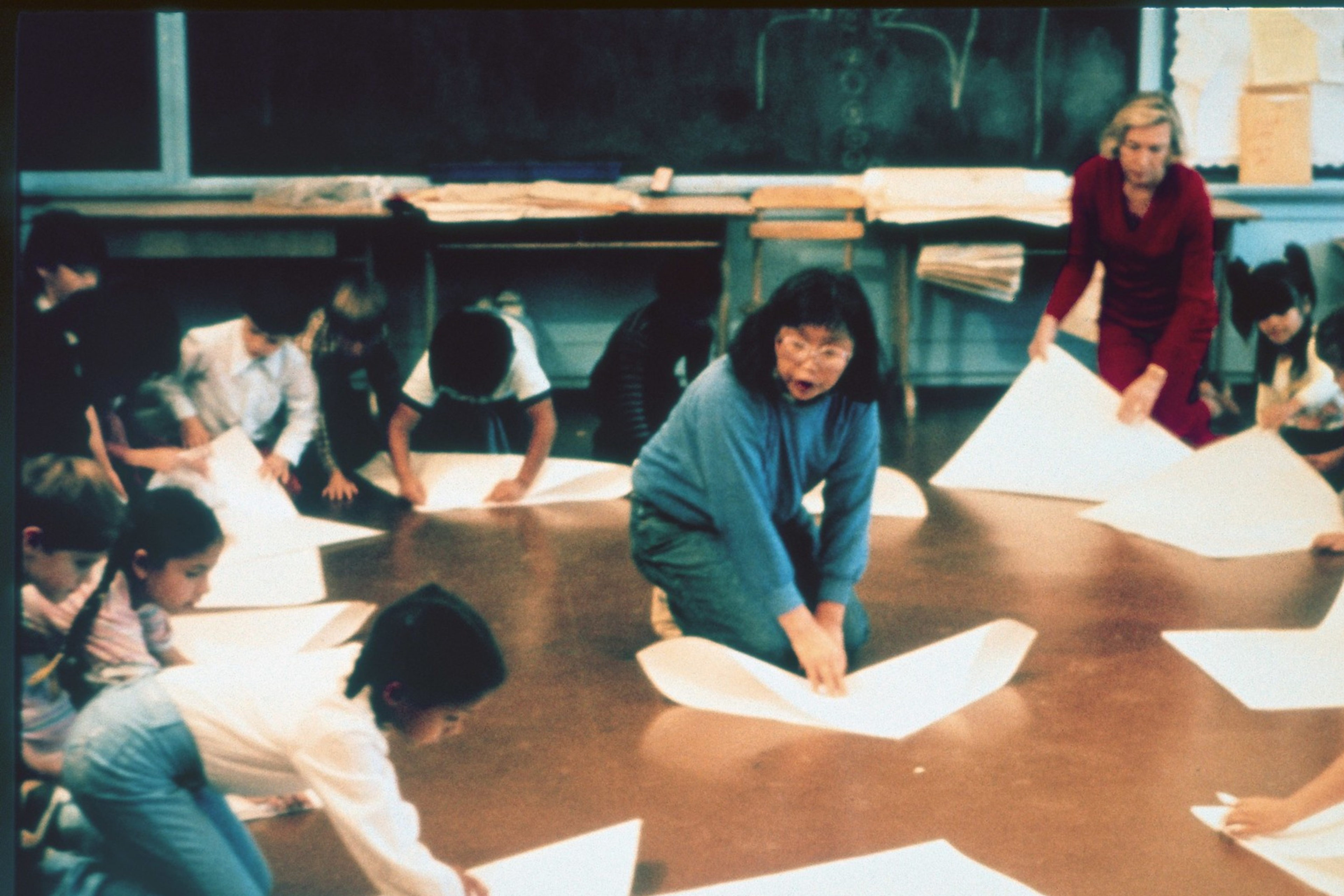 A group of children and two adults are gathered in a circle on the floor, engaged in folding large sheets of paper, in a classroom setting.
