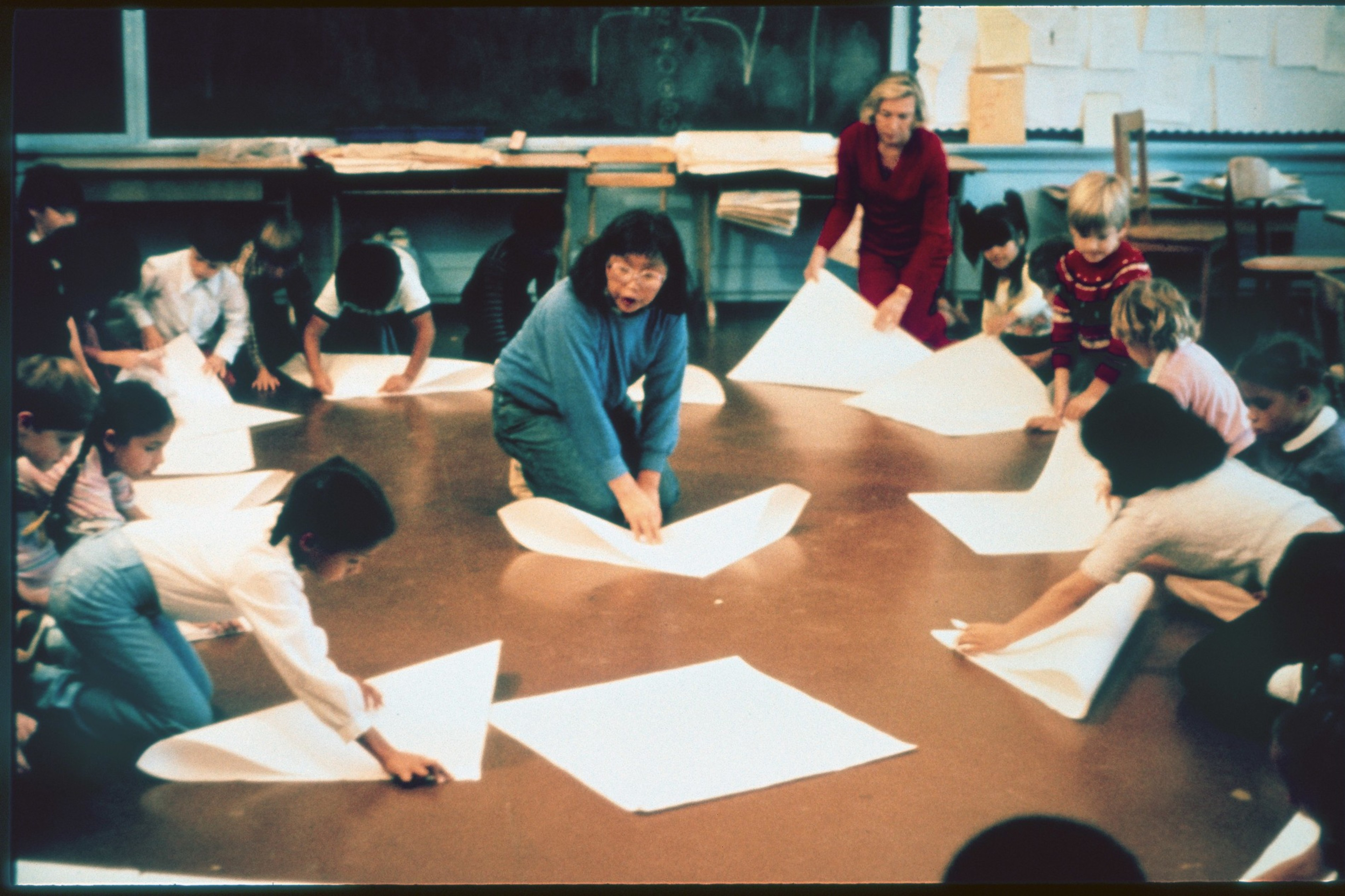 A group of children and two adults are gathered in a circle on the floor, engaged in folding large sheets of paper, in a classroom setting.