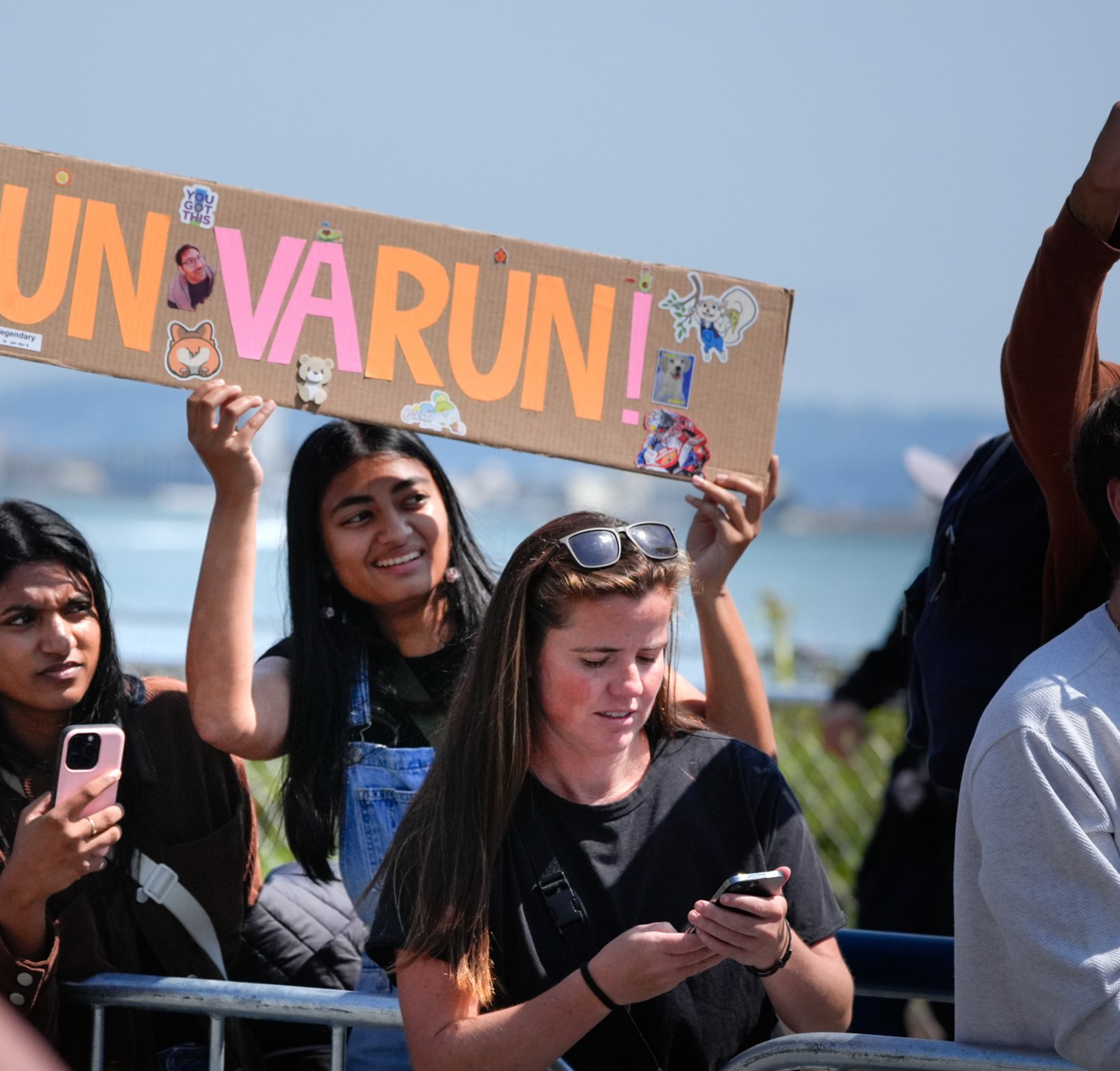 A group of people are watching an event outdoors, with one person holding a sign that says &quot;RUN VARUN RUN!&quot; while another looks at their phone.