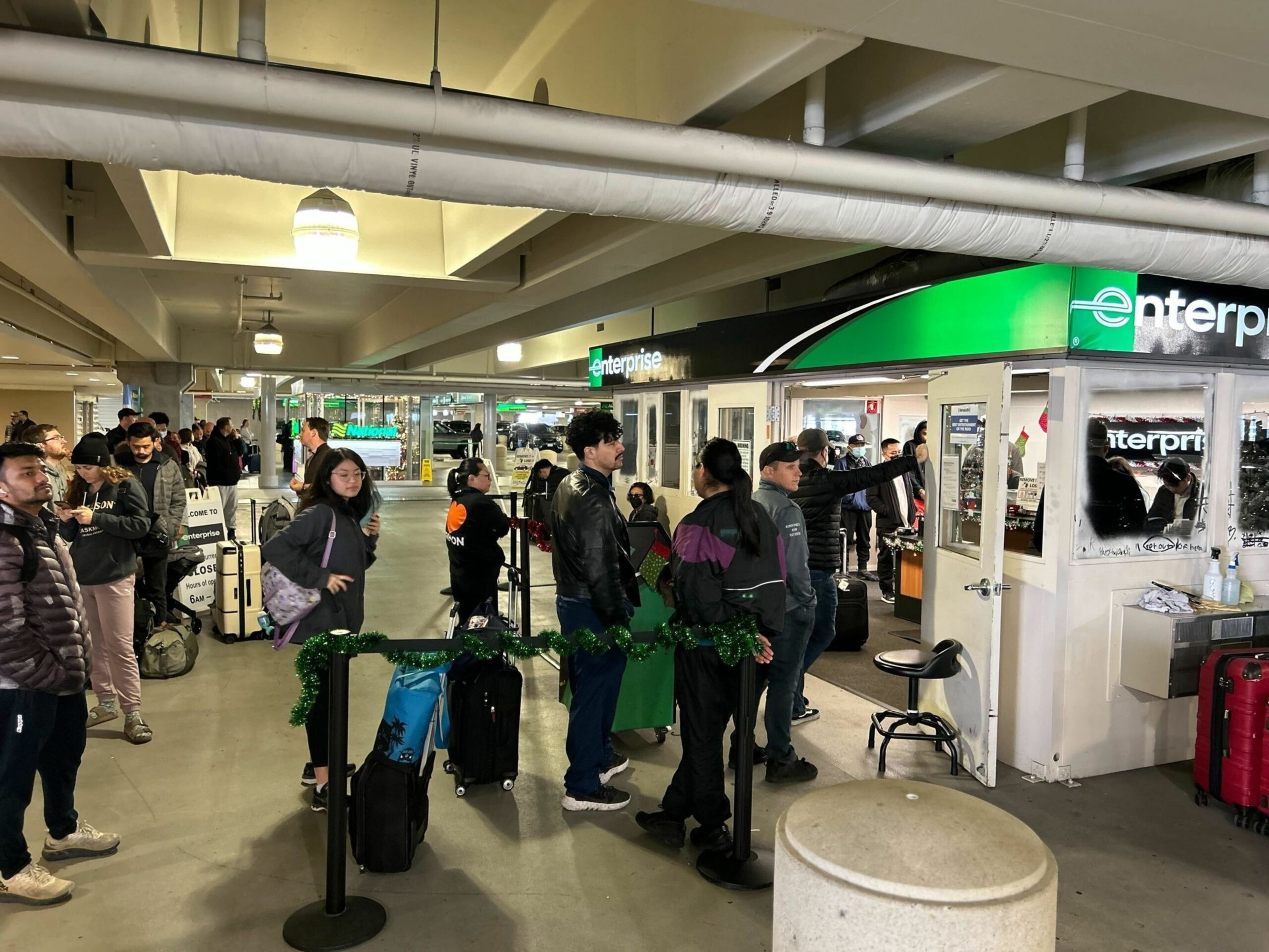 People stand in line with suitcases and bags inside a parking garage, waiting at an Enterprise car rental counter decorated with green garland.
