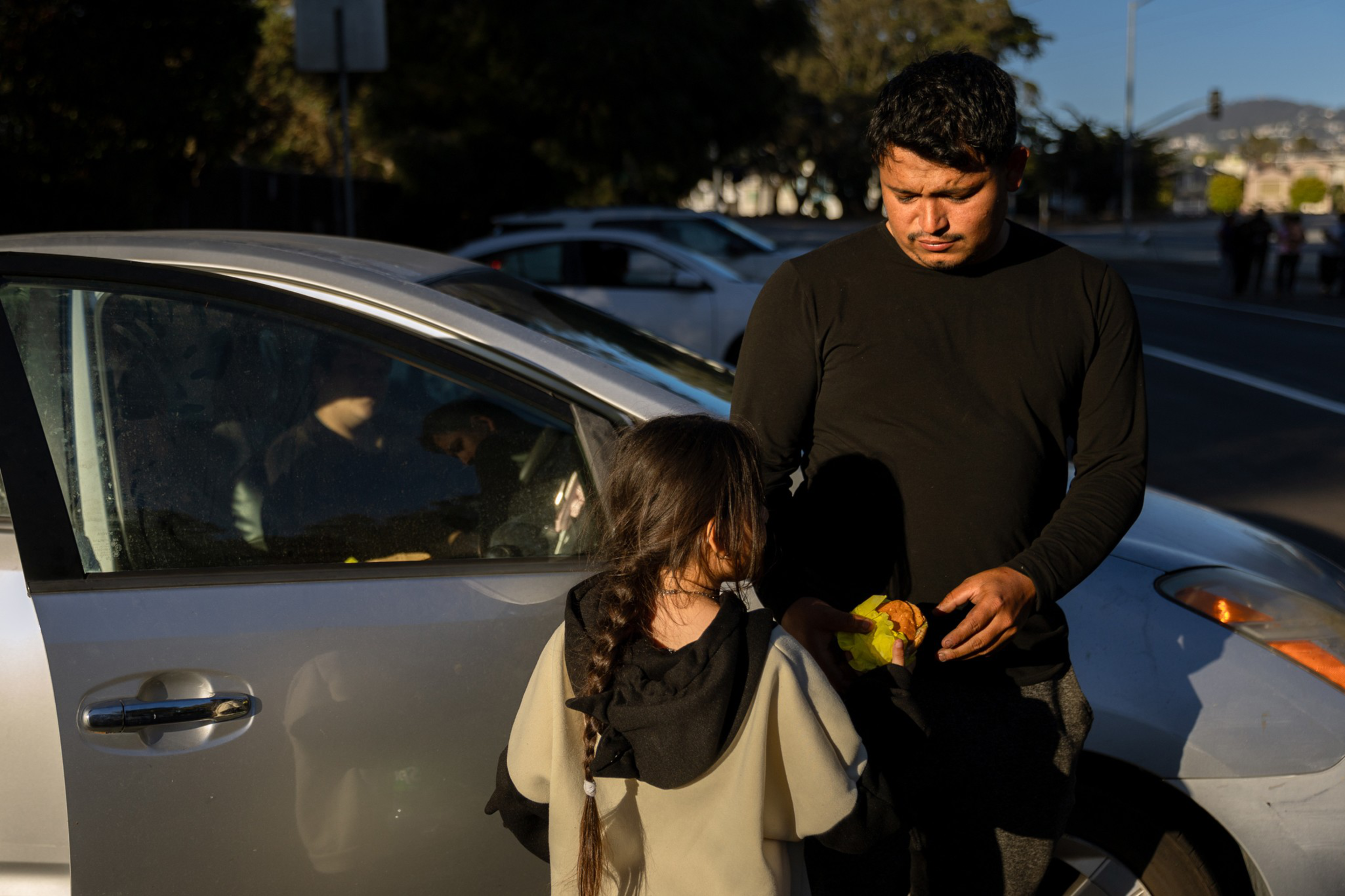 A man in a black shirt holds a burger while standing by a silver car, facing a child with a braid. The car door is open, and the scene is set on a sunny day.