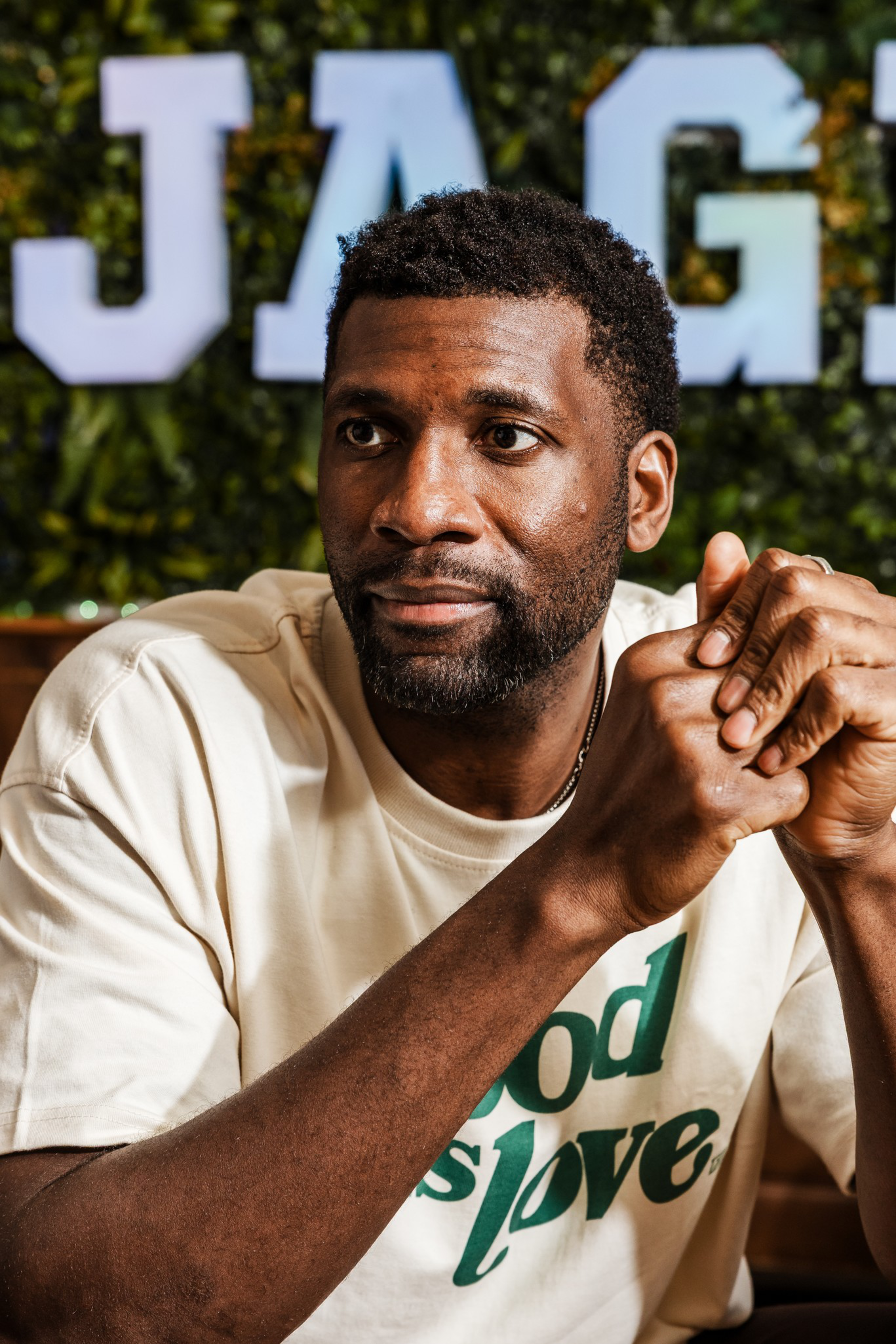 A man with short curly hair and a beard clasps his hands, wearing a cream shirt with green text and sitting in front of a green leafy background with large letters.