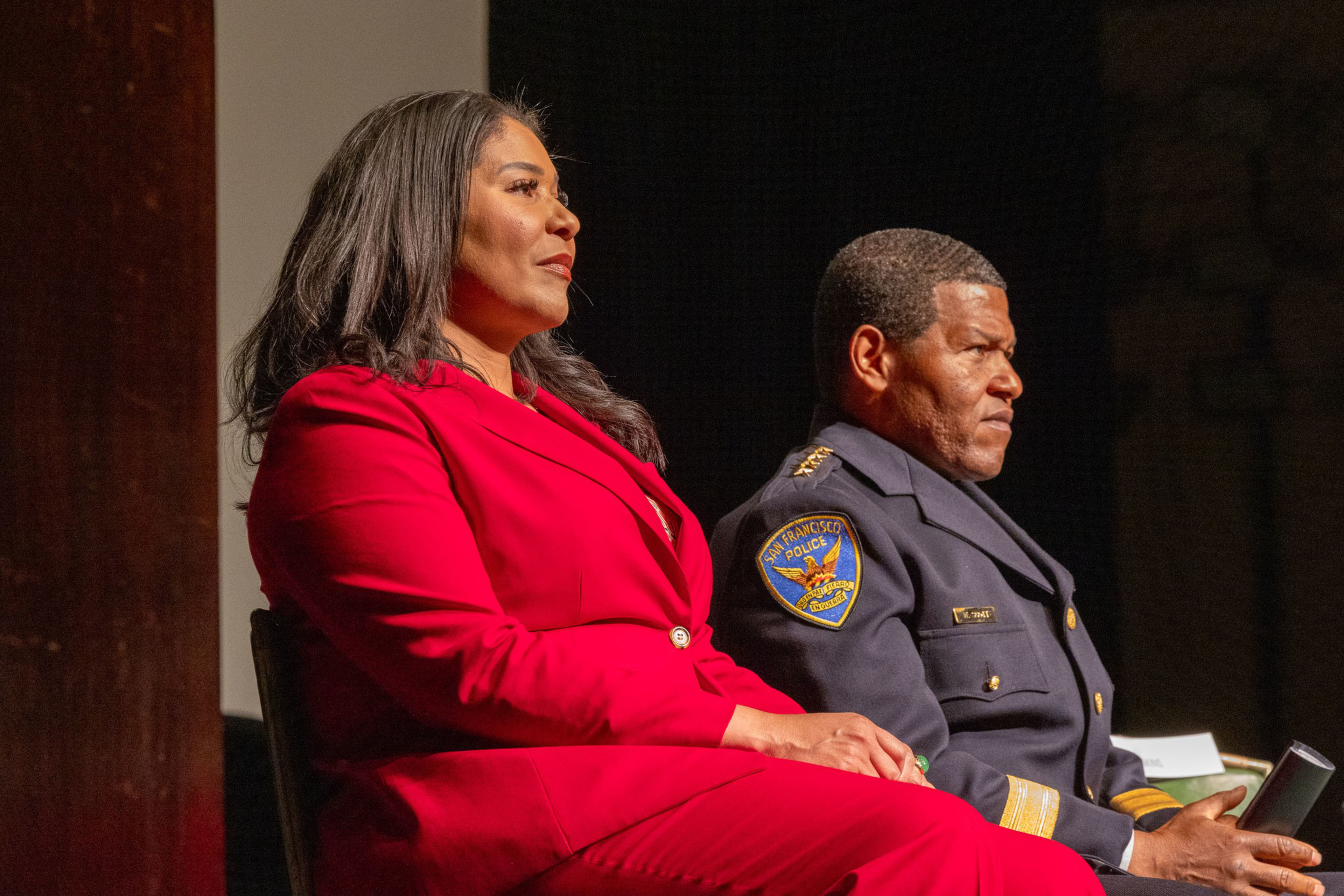 A woman in a red suit and a man in a San Francisco police uniform sit side by side, both looking forward with serious expressions.