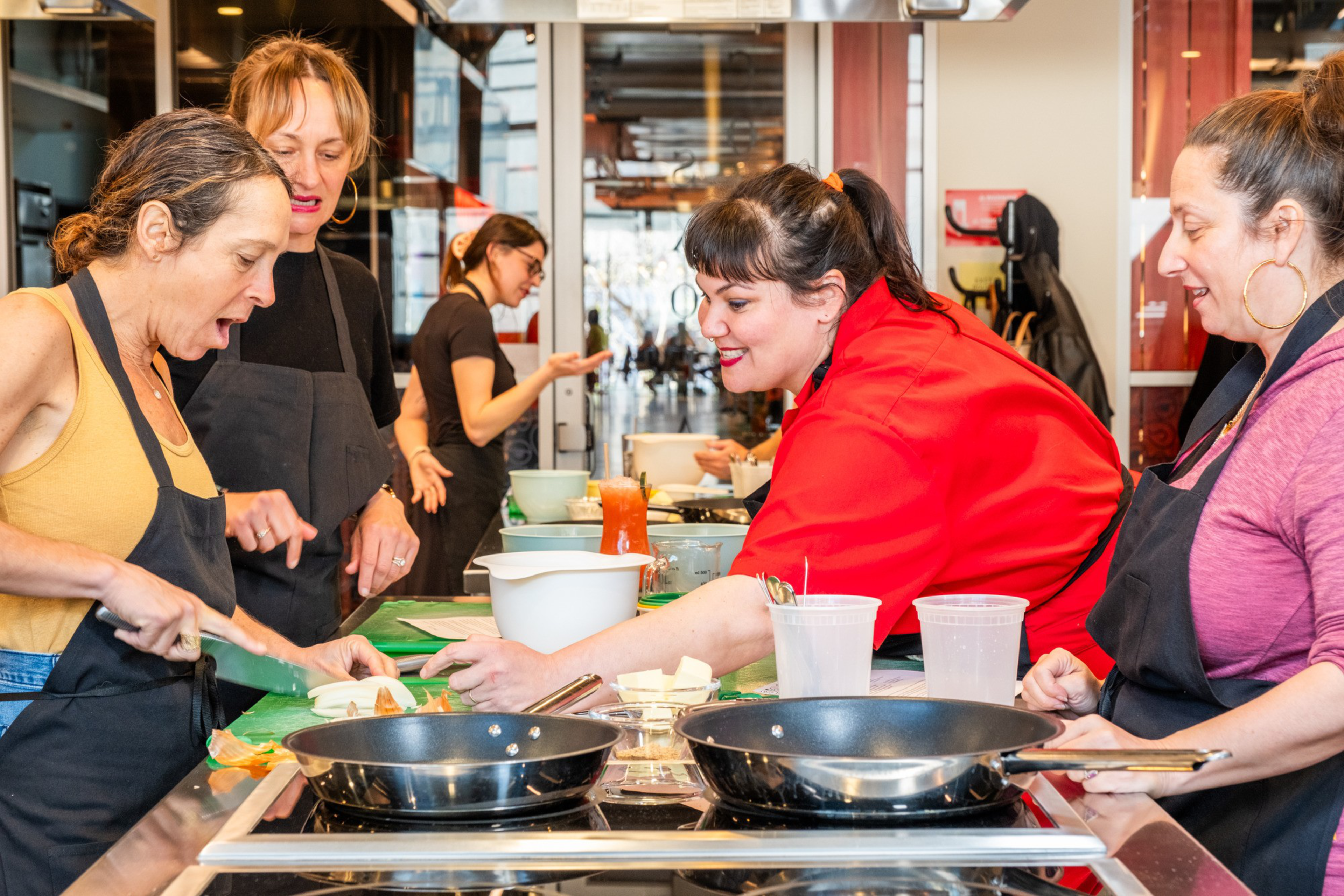 Four people in a kitchen wear aprons, focusing on chopping vegetables. A chef, dressed in red, observes. Cooking utensils and ingredients fill the counter.