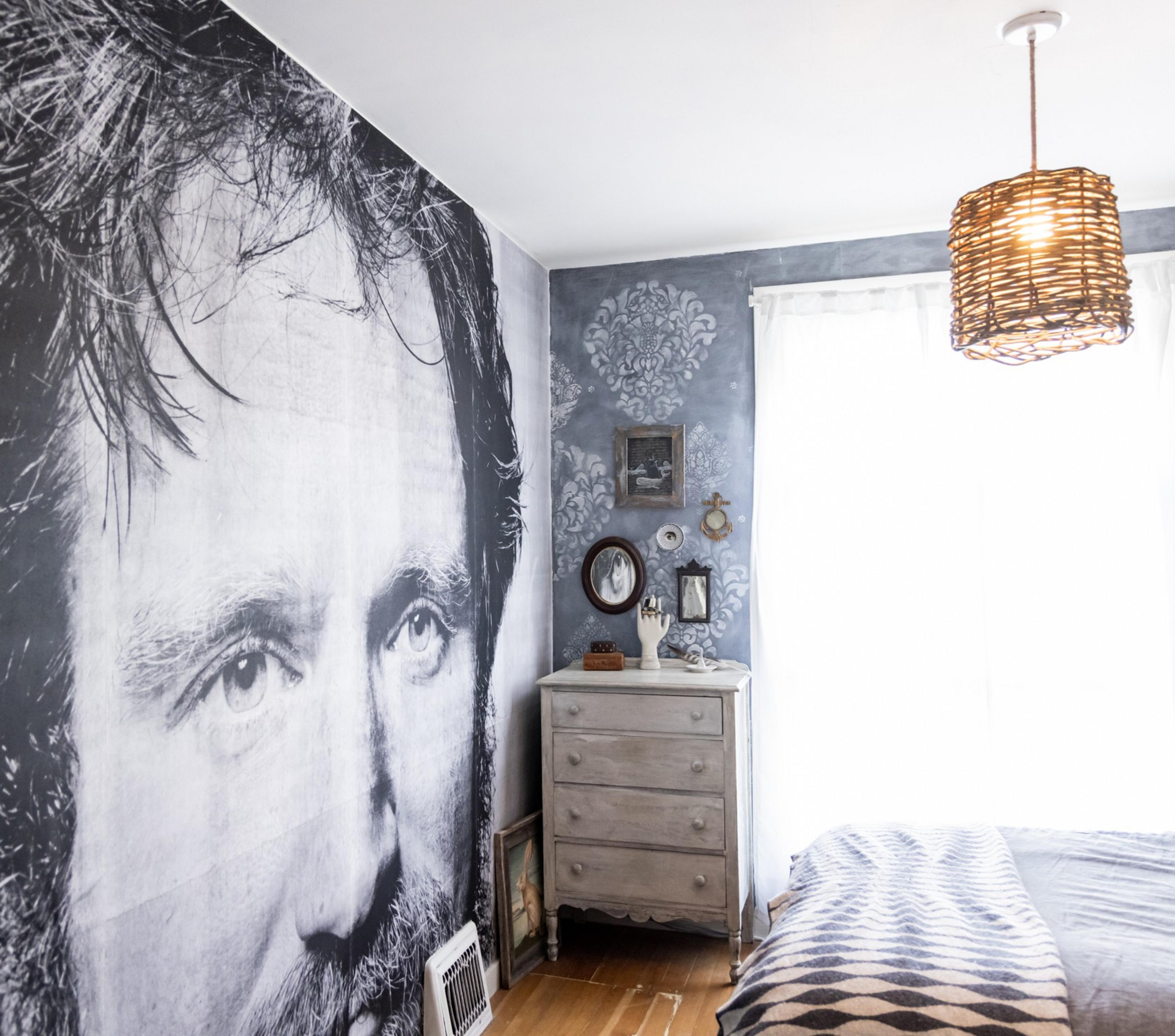 The image shows a bedroom with a large grayscale mural of a man's face on the wall. There's a dresser with mirrors, decorative items, and a woven hanging light.