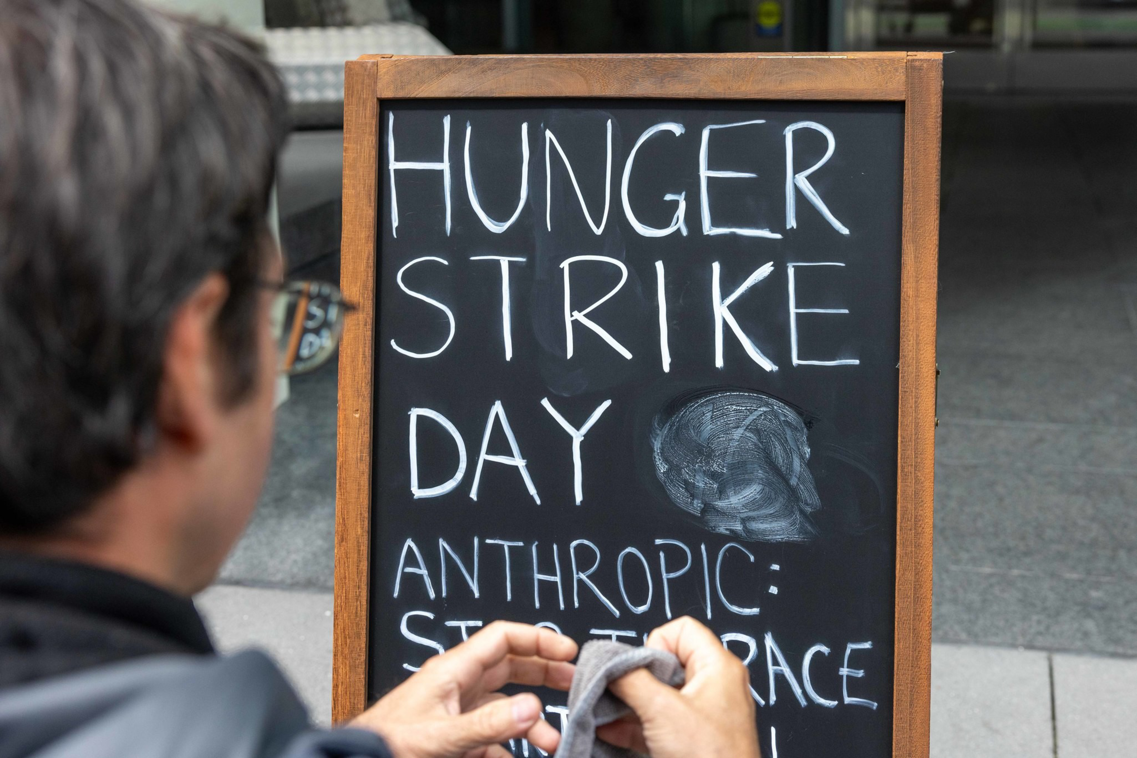 A person cleans a blackboard sign that reads “Hunger Strike Day” with additional text below that is partially erased.