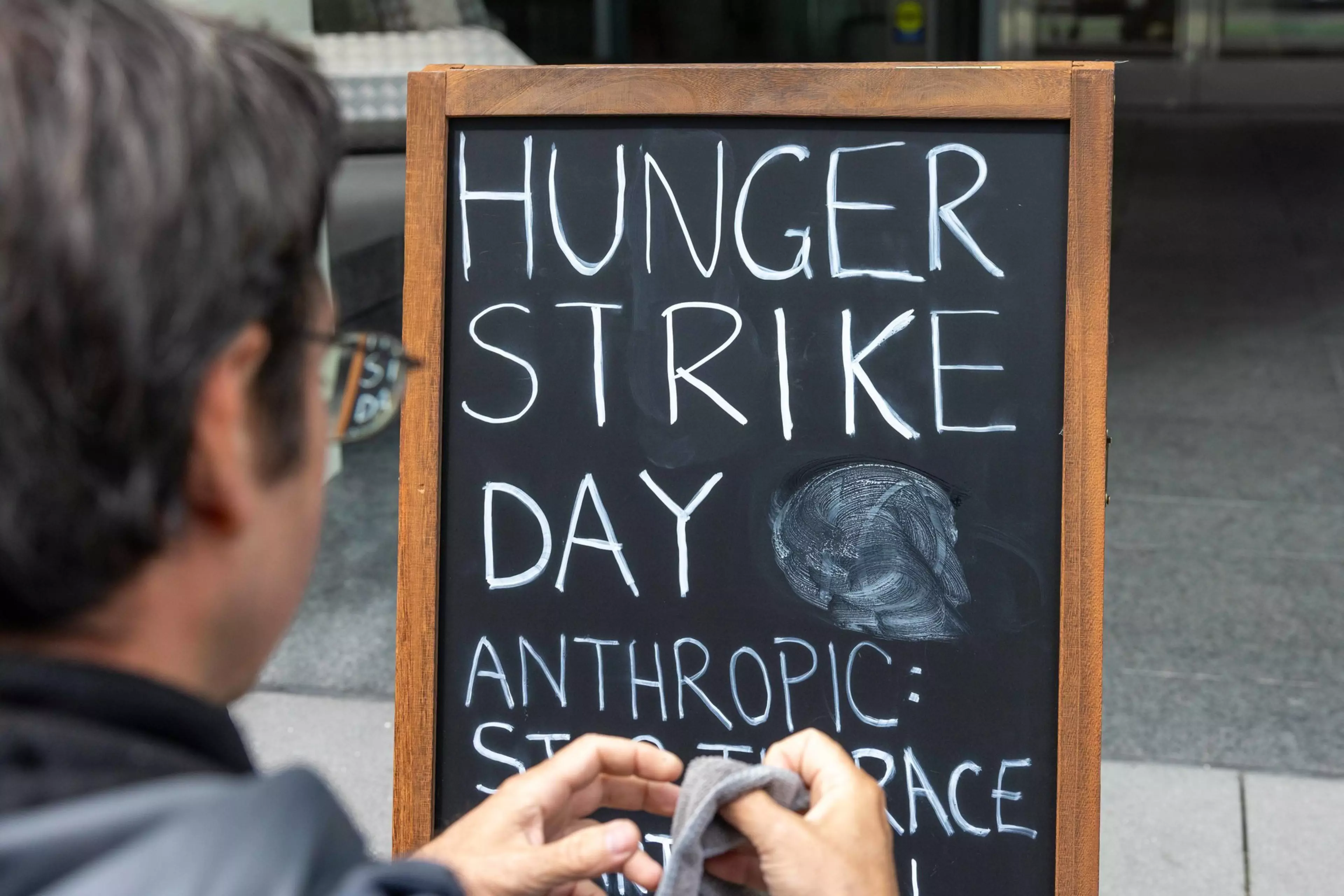 A person cleans a blackboard sign that reads “Hunger Strike Day” with additional text below that is partially erased.