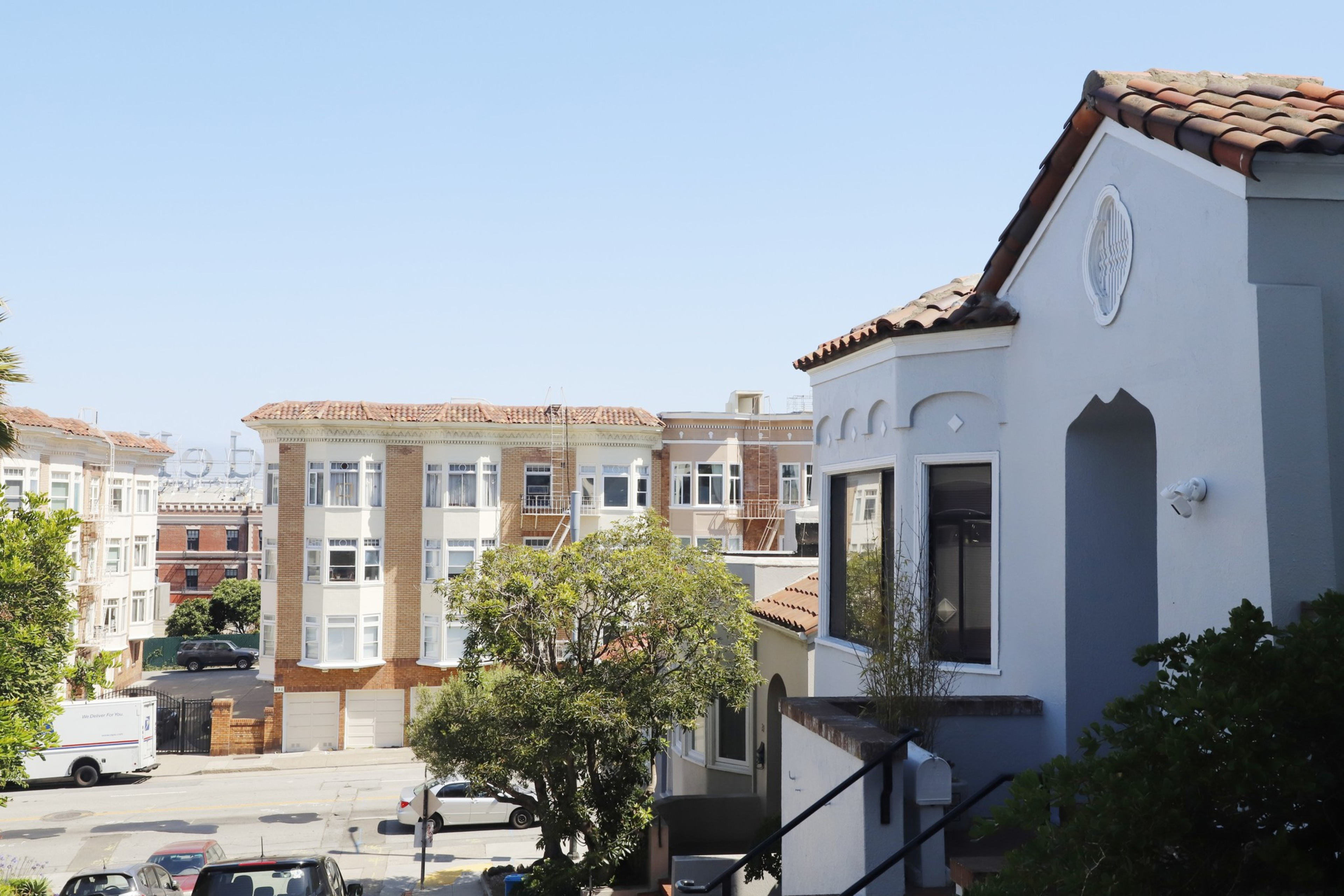 The image shows a street lined with tan, multi-story buildings featuring multiple large windows. There are trees, cars, and a white truck on the street.