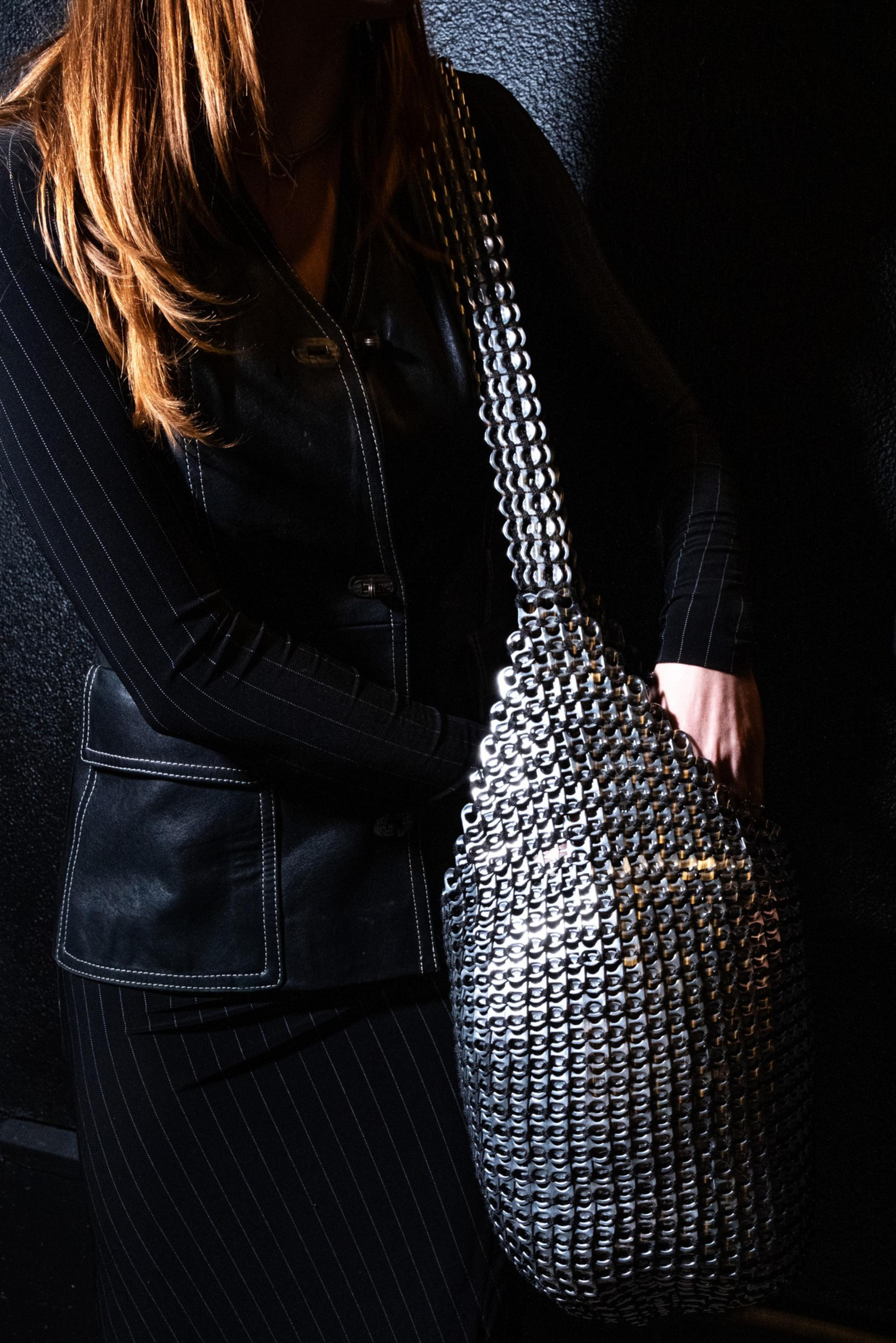 A person in a black pinstripe outfit holds a shiny, metallic handbag made of interlinked silver tabs, against a dark textured background.