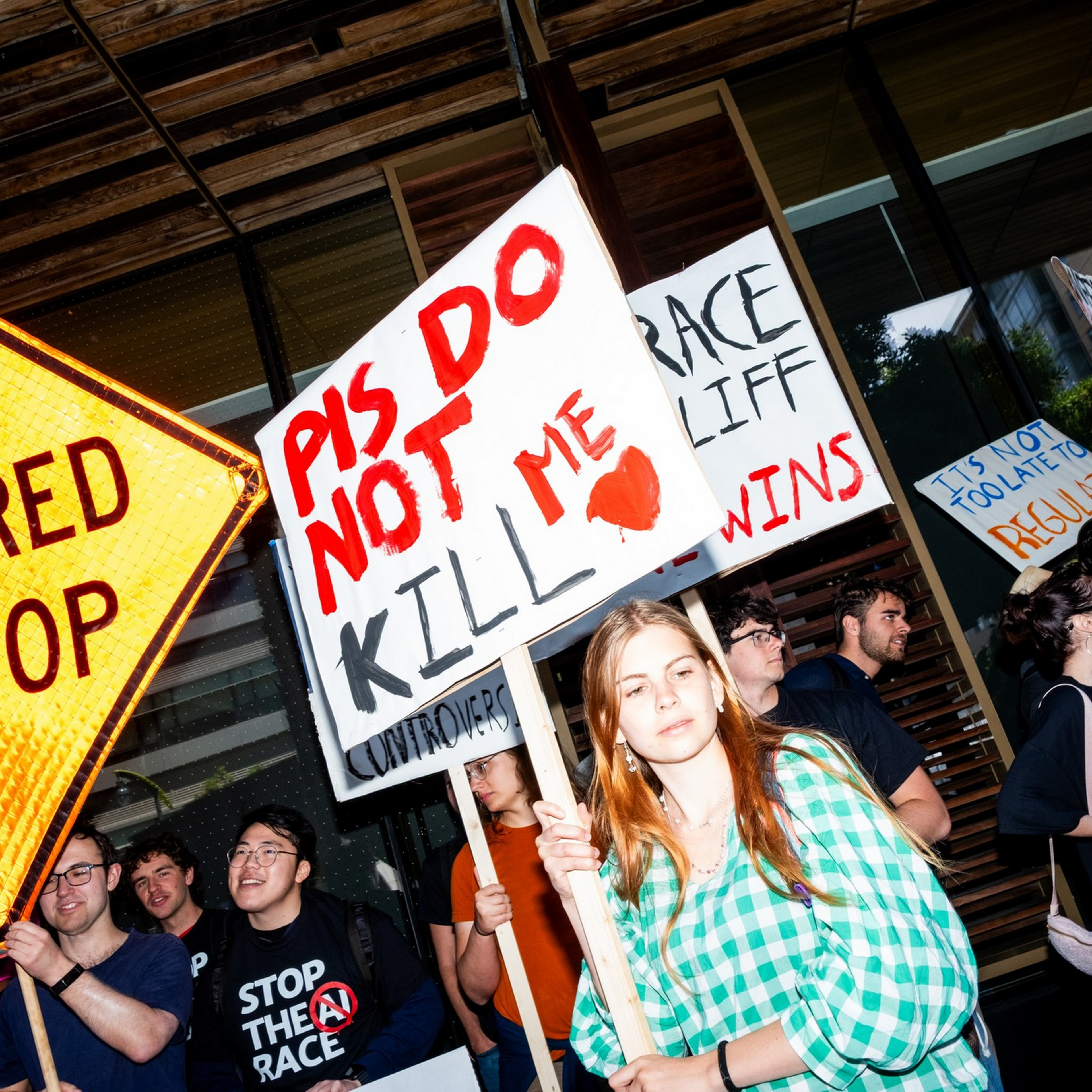 People hold protest signs including "PIS DO NOT KILL ME" and "BE PREPARED TO STOP" outside a modern building during a daytime rally.