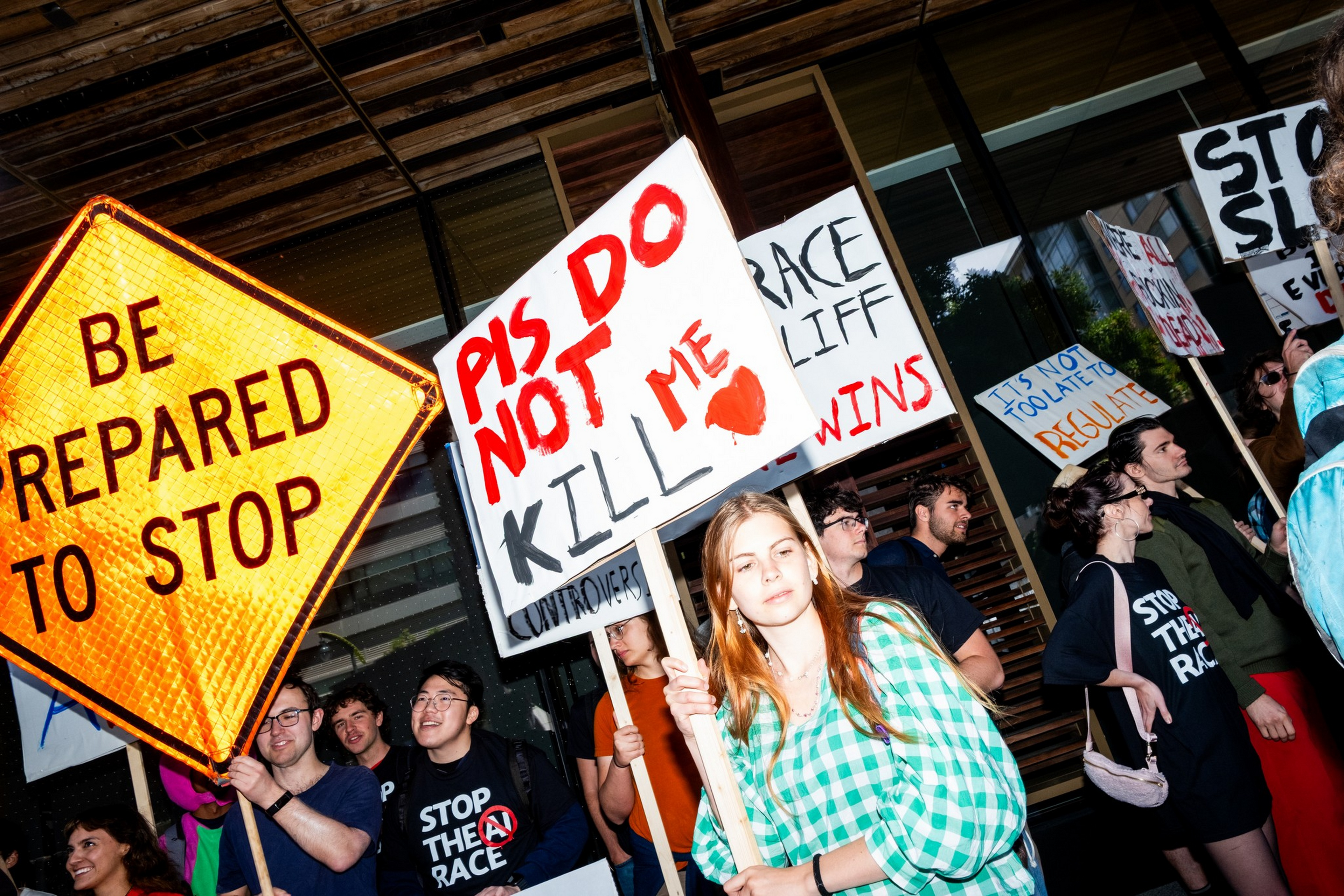 People hold protest signs including "PIS DO NOT KILL ME" and "BE PREPARED TO STOP" outside a modern building during a daytime rally.