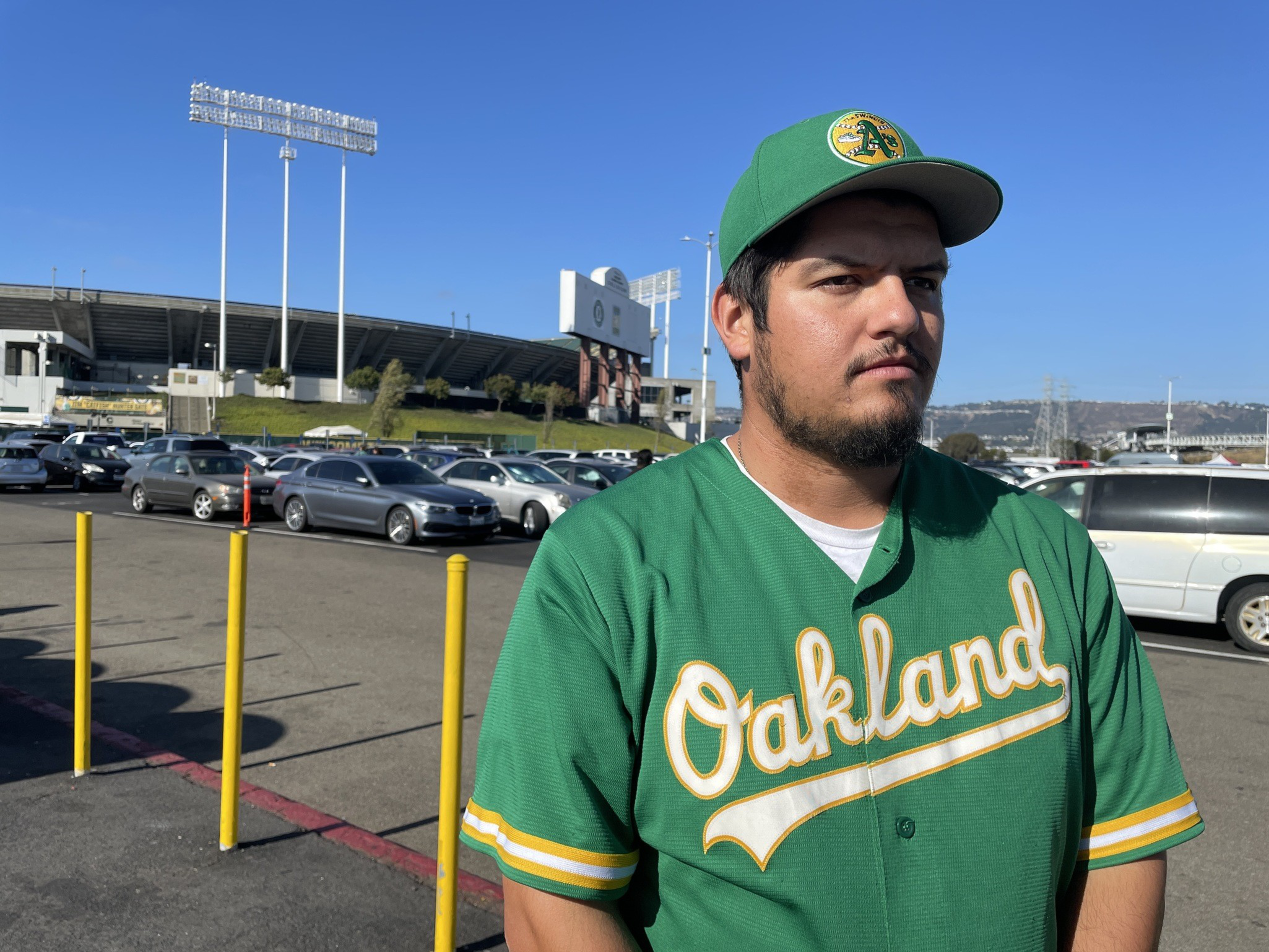 A man in a green baseball jersey and cap stands in a parking lot with a large stadium visible in the background under a clear blue sky.
