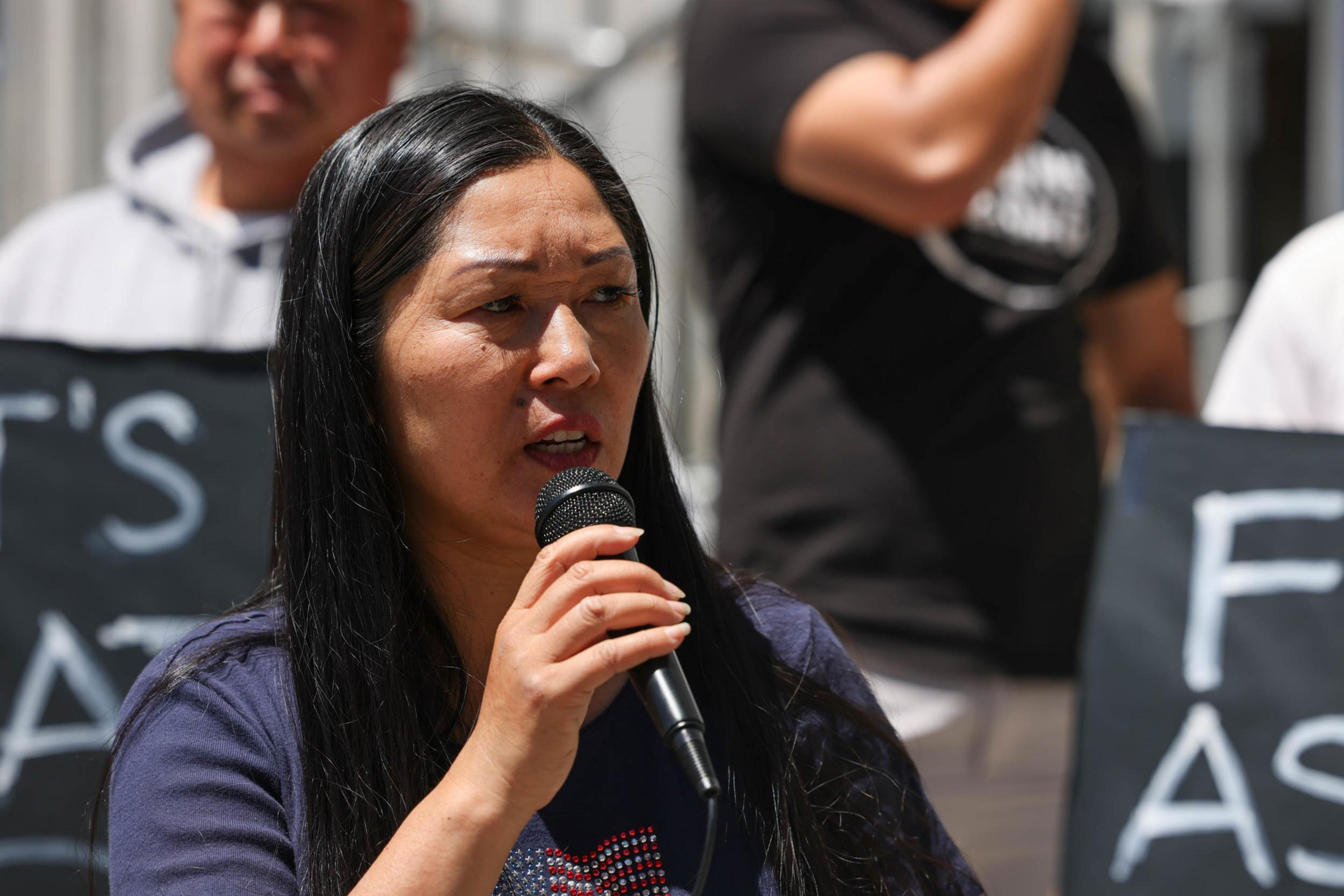 A woman with long black hair holds a microphone and speaks, with two blurred people and part of protest signs visible in the background.