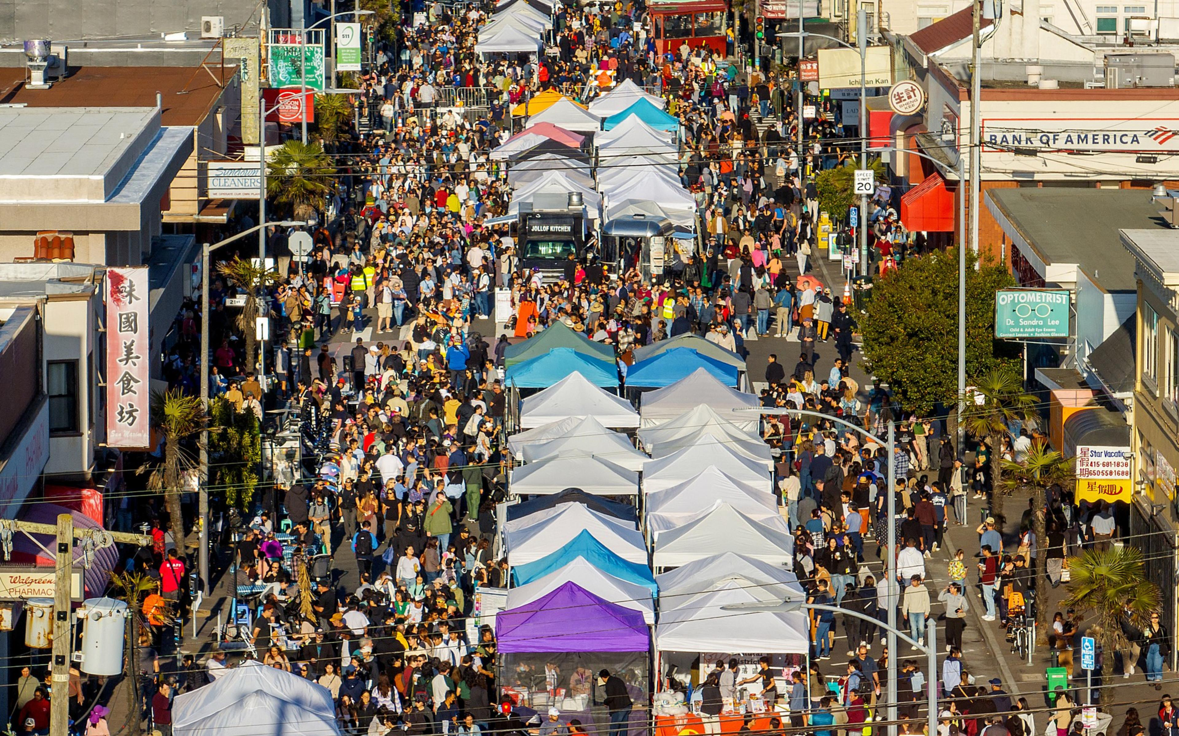 A bustling street fair with numerous white tents and a large crowd of people. The lively scene includes colorful booths and various storefronts on both sides.