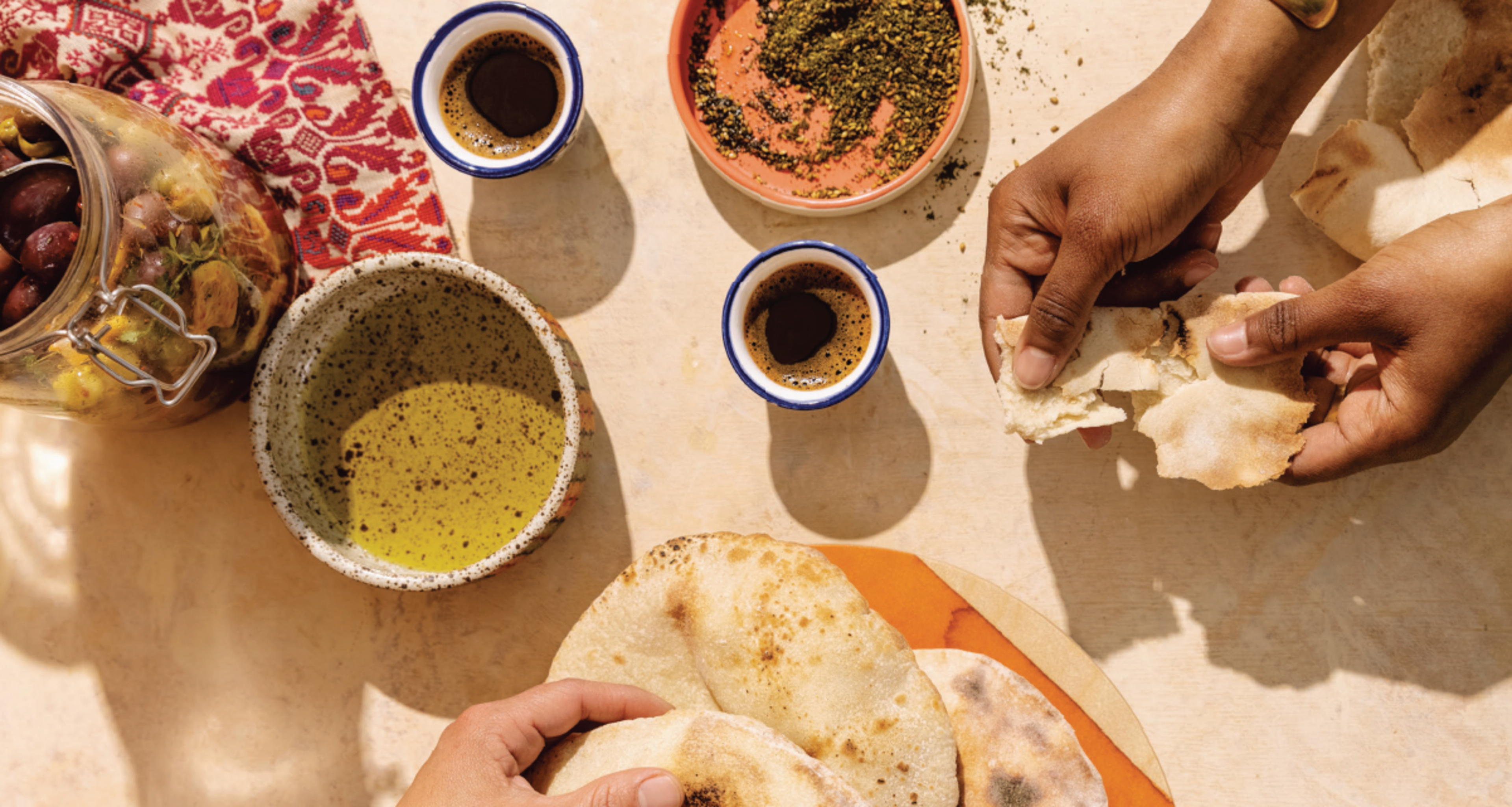Hands are breaking flatbread near bowls of olive oil, za'atar spice, two cups of espresso, and a jar of olives on a light-colored table.