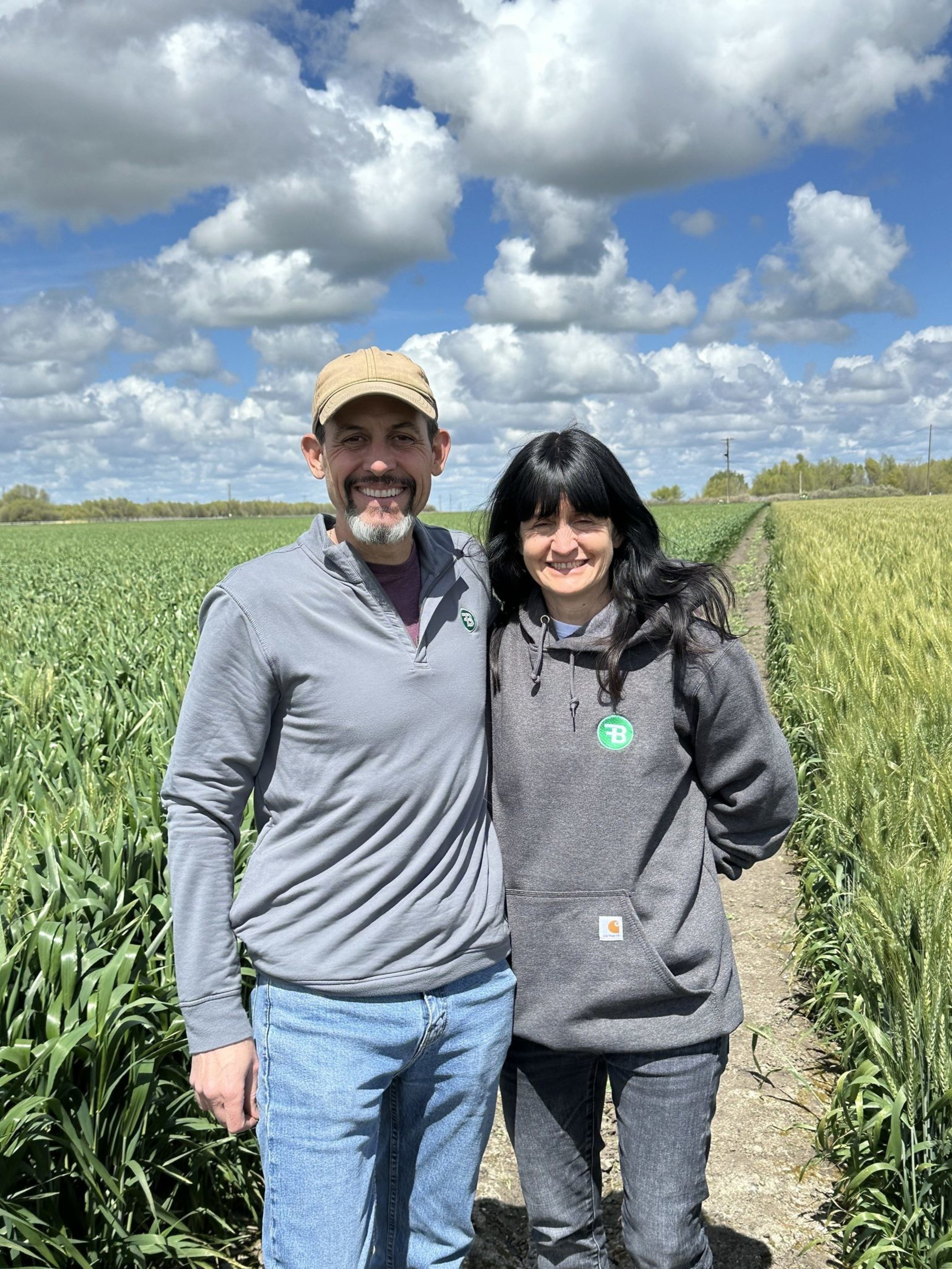 A smiling couple stands side by side on a path through lush green fields, beneath a sky filled with fluffy clouds. Both wear casual clothes and look content.