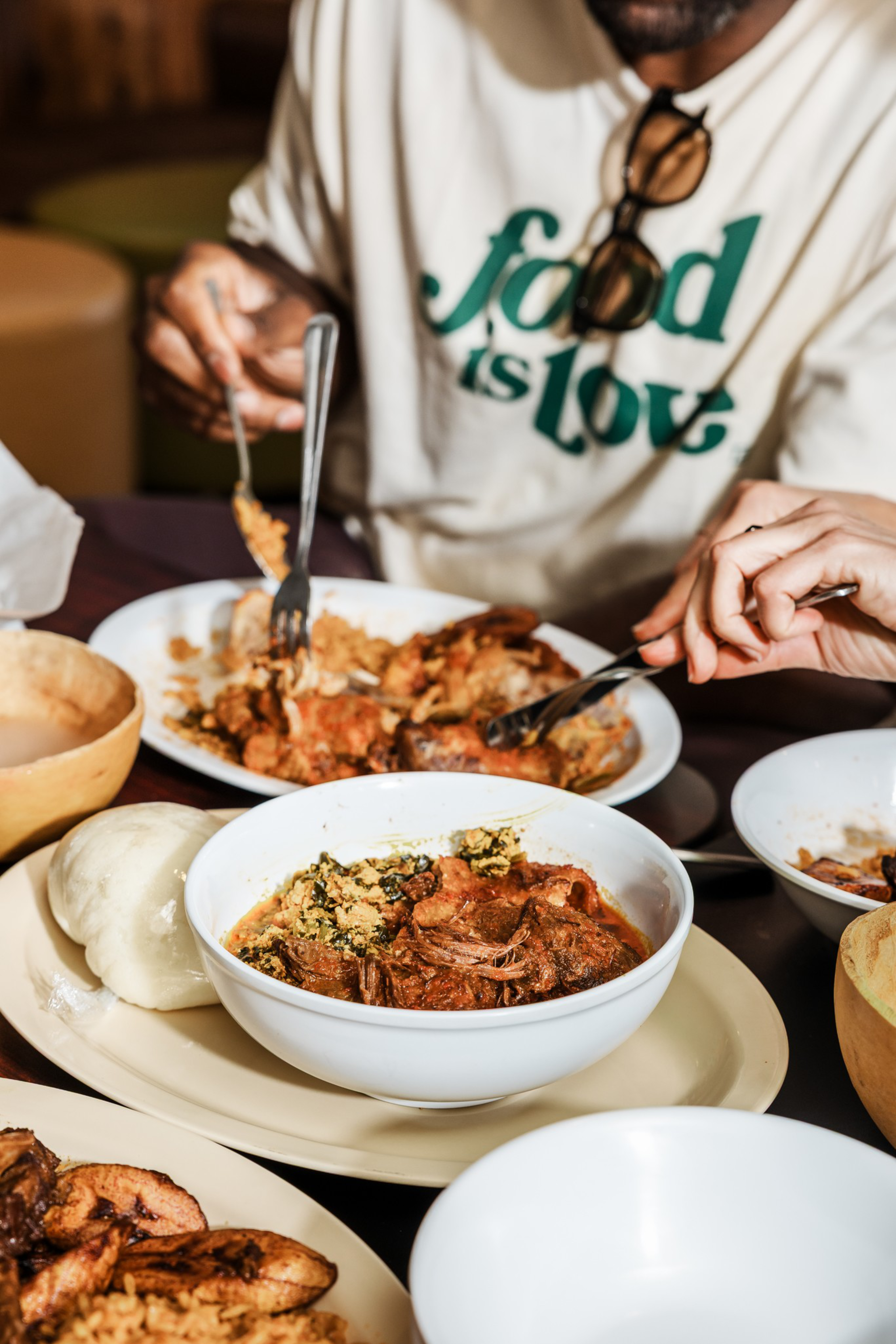 Two people are sharing a meal with bowls of meat stew, fufu, fried plantains, and other dishes on a table.