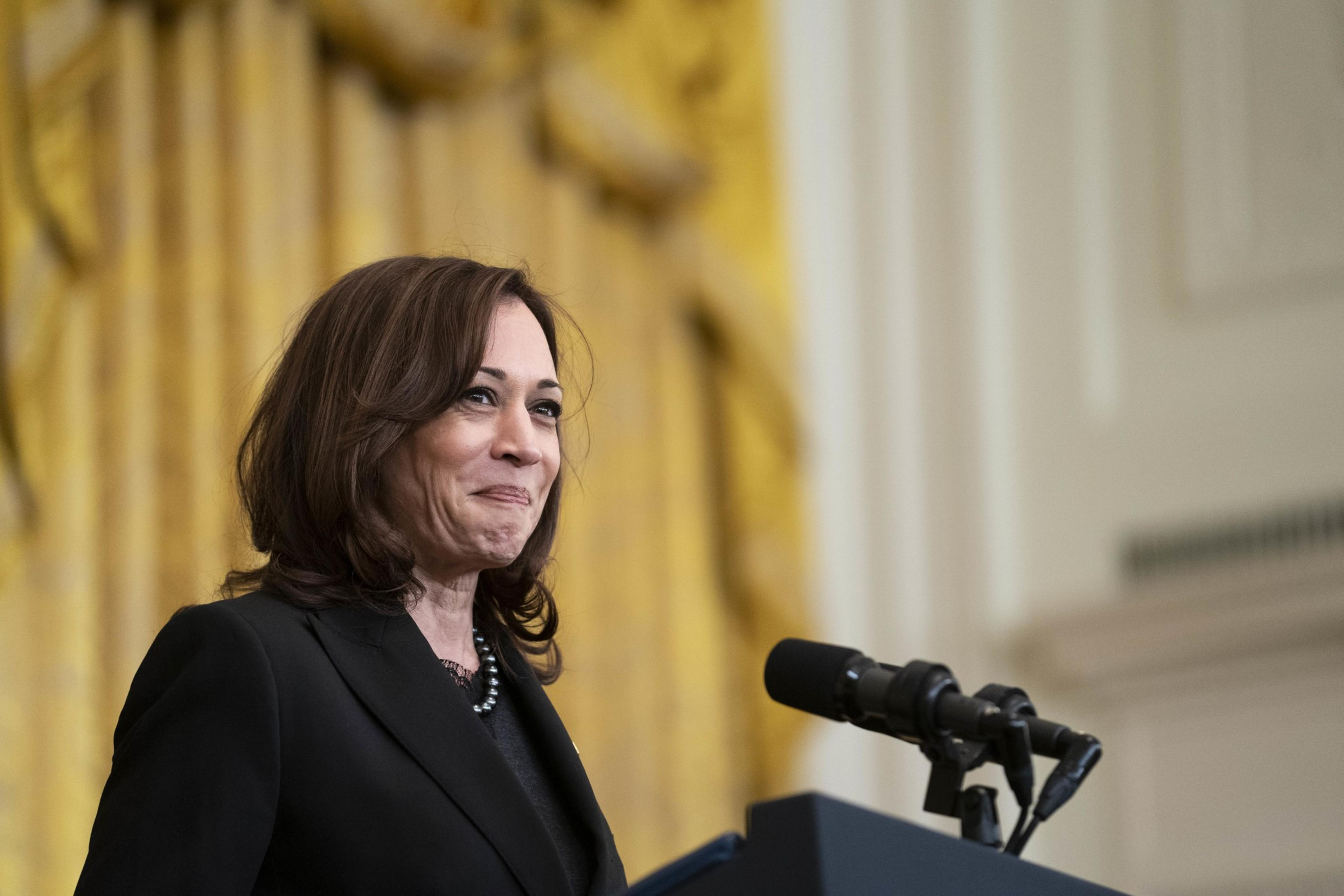 A woman wearing a black suit stands at a podium with microphones in front of a golden curtain, smiling slightly.