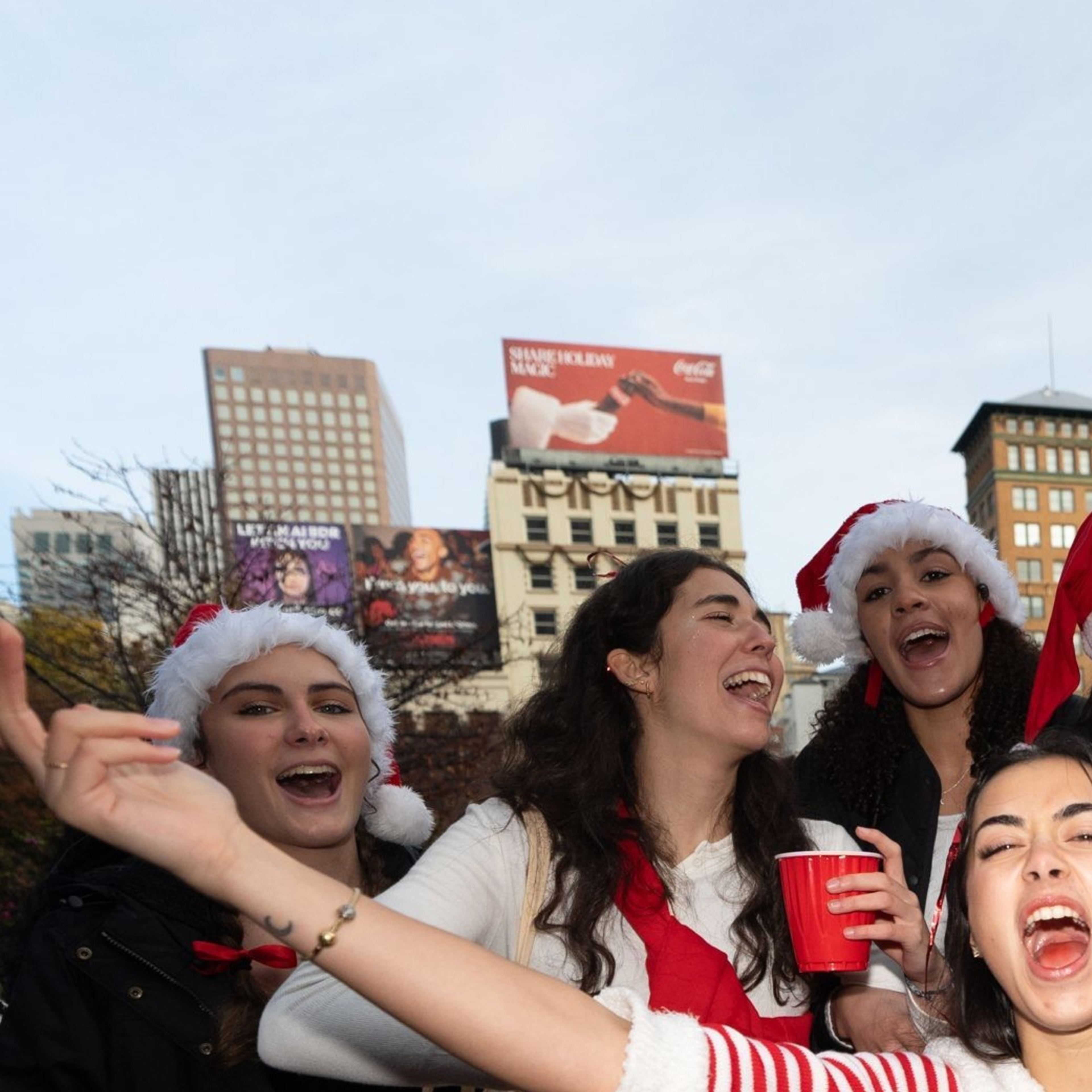 A joyful group of people, mostly women wearing festive Santa hats and red outfits, are celebrating outdoors near a decorated Christmas tree.