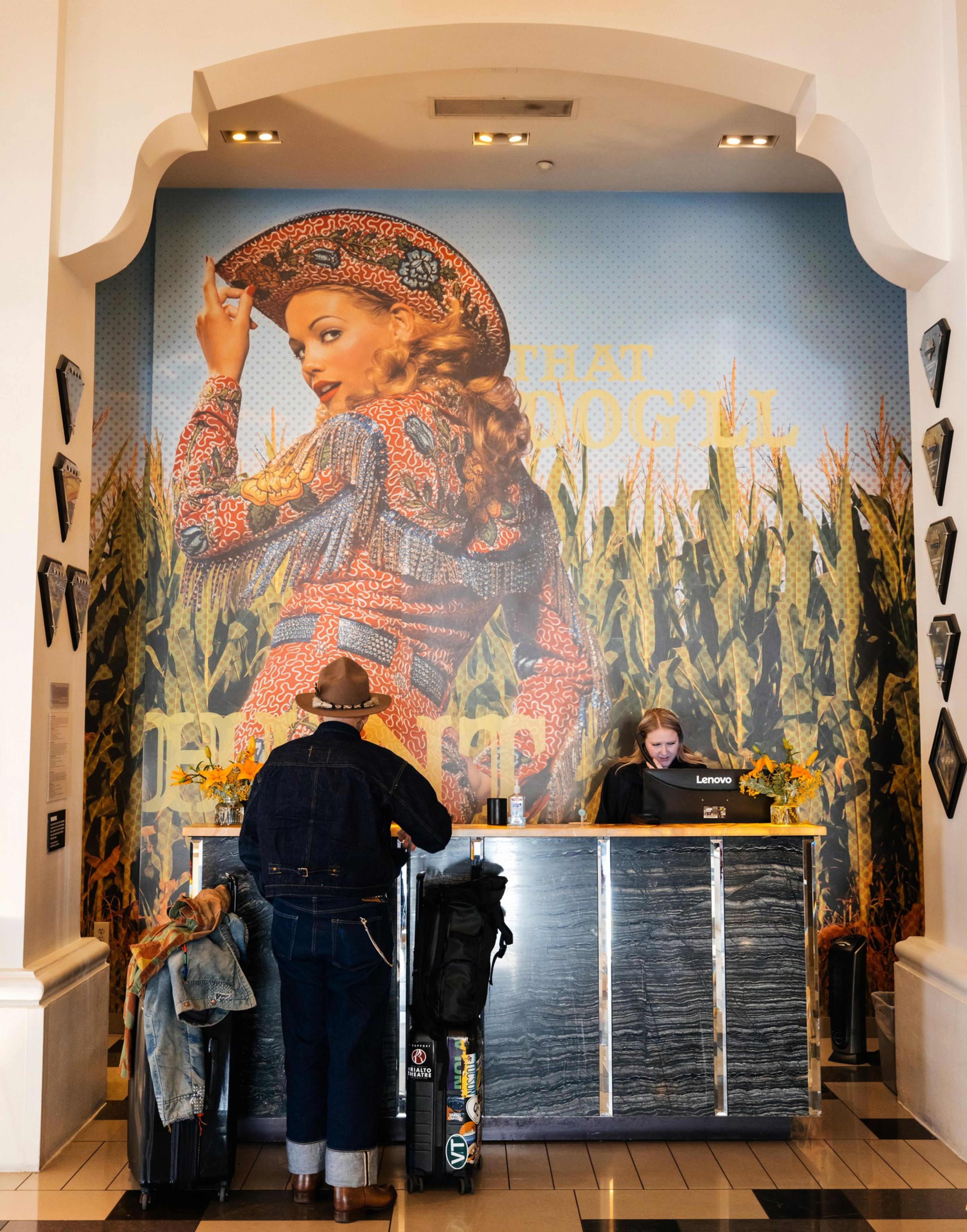 A man in denim stands at a reception desk with luggage. Behind him, a large mural of a woman in a cowboy hat is displayed, with cornfields in the background.