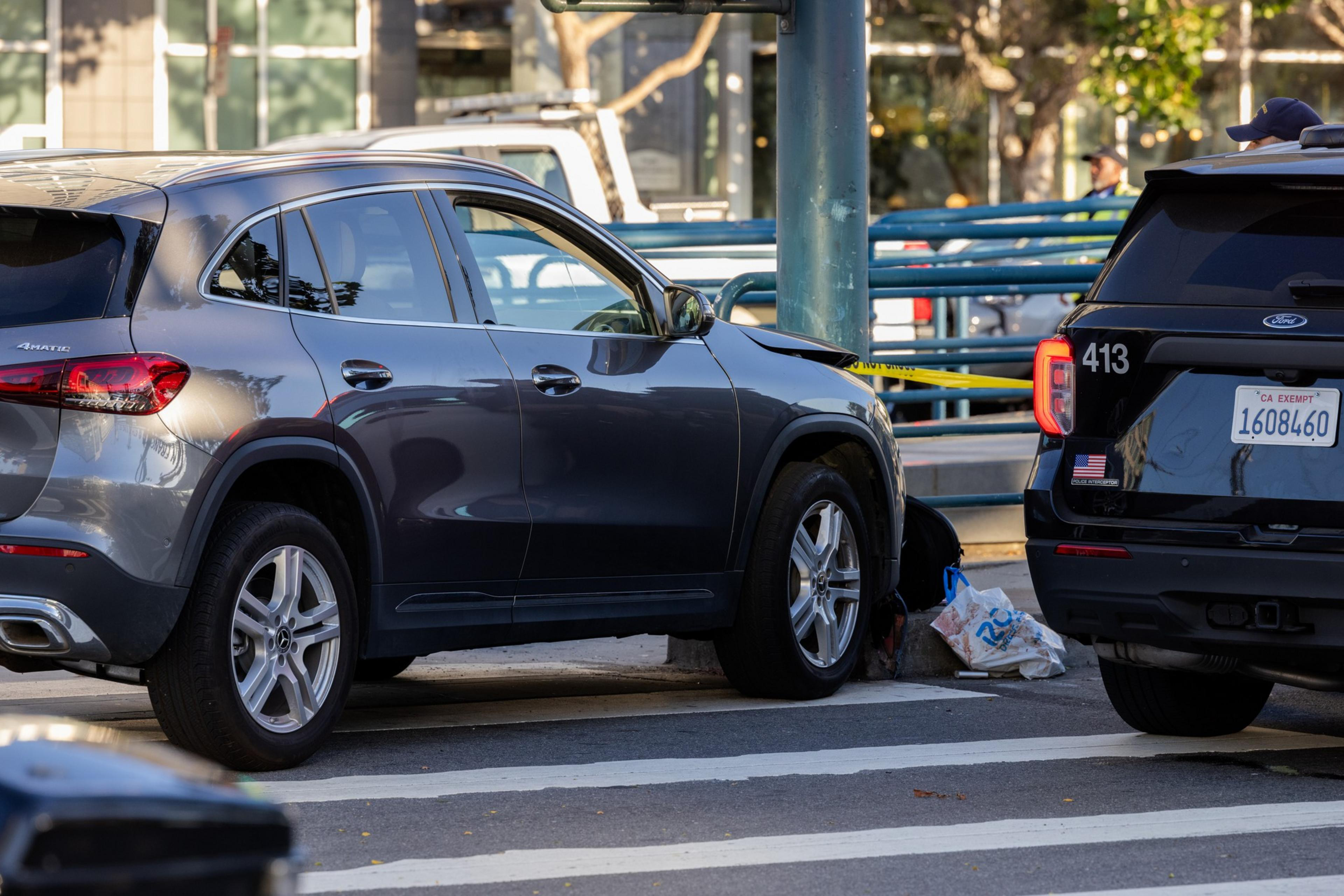 A gray SUV is parked next to a black police car with yellow caution tape around the scene and a person crouched near the SUV's front tire.