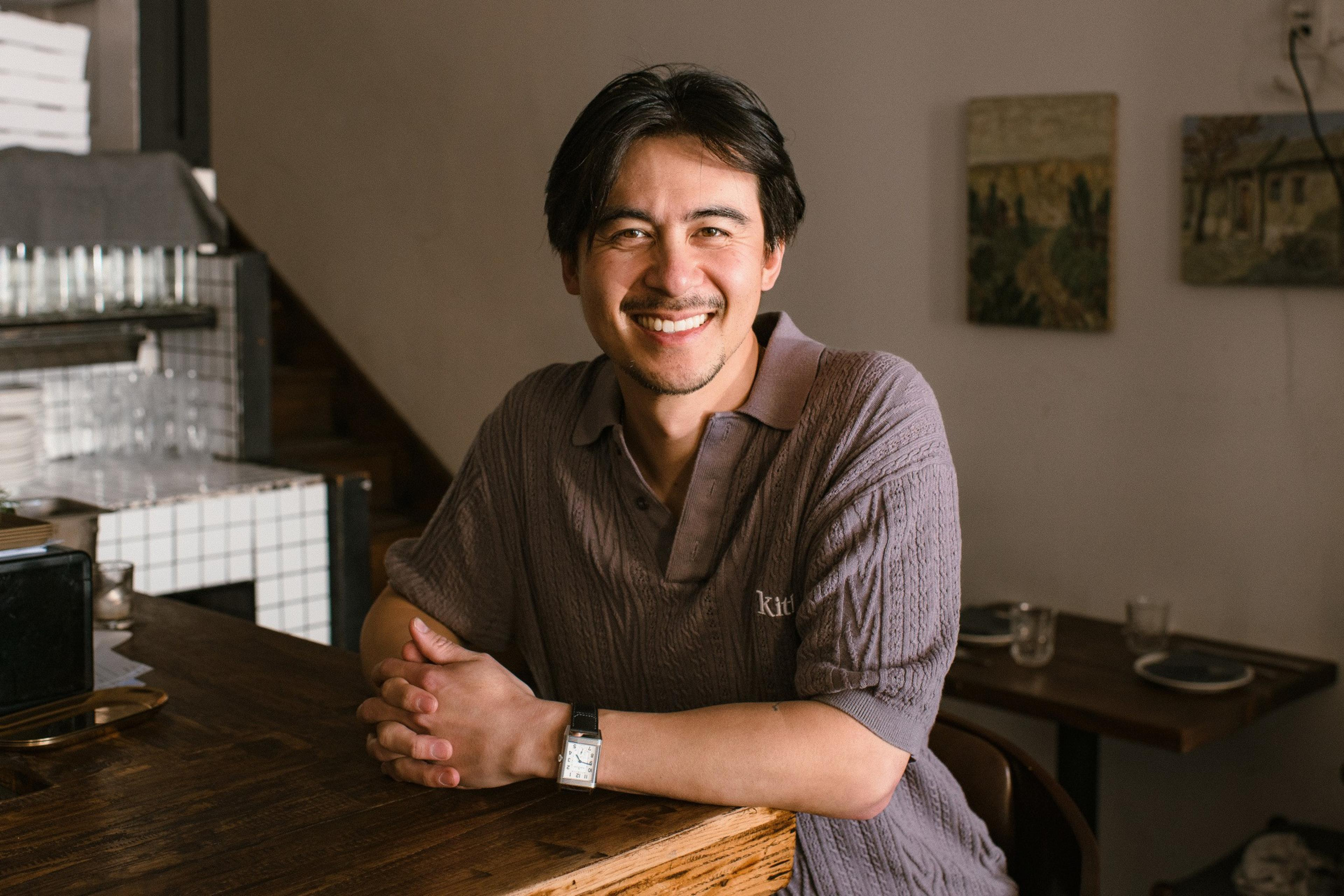 A person with a friendly smile sits at a wooden table in a cozy room, wearing a gray shirt. The background includes a shelf and framed artwork.
