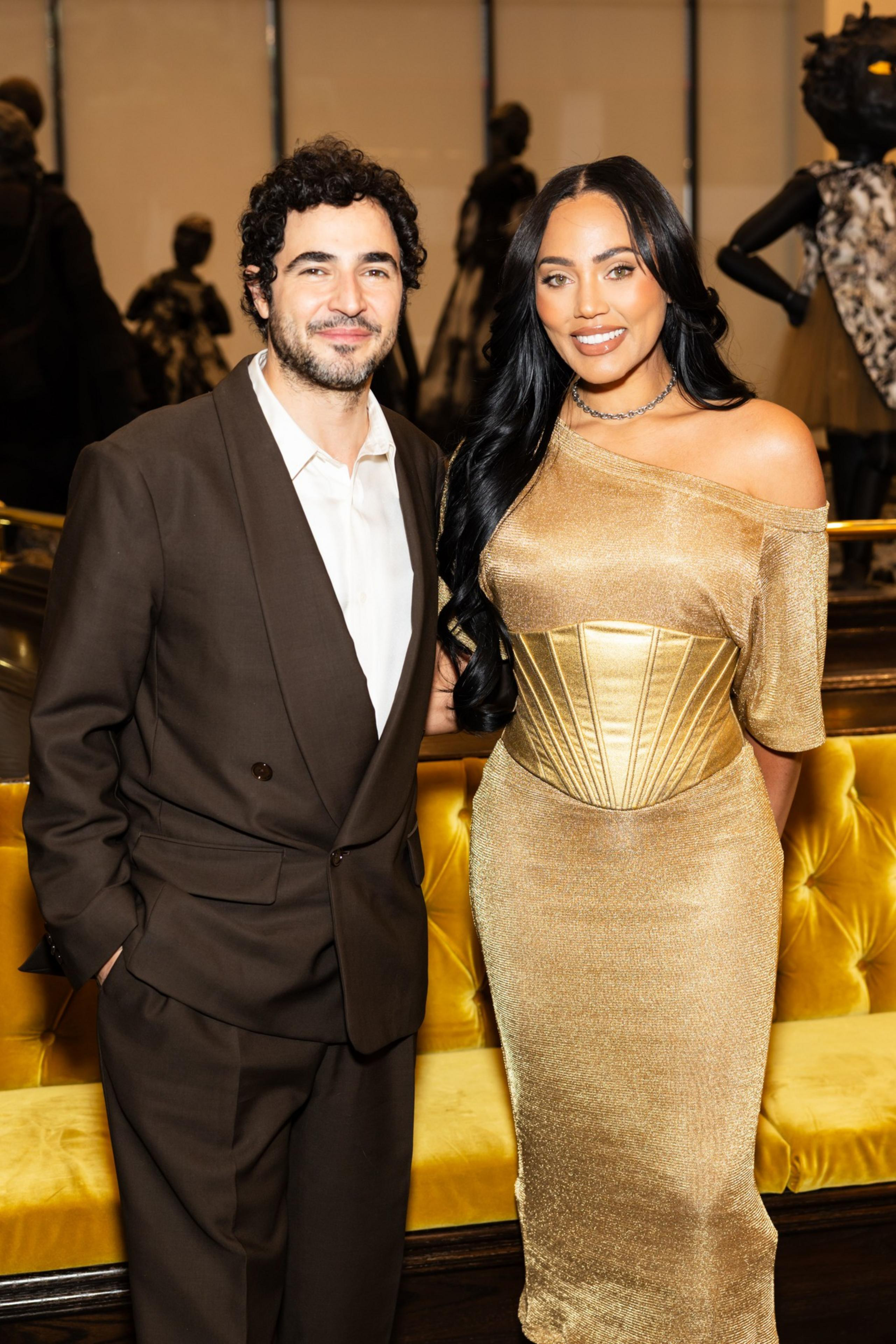 A man in a brown suit and open-collar white shirt stands next to a woman in a gold gown with an off-the-shoulder design. They are in front of a yellow couch.