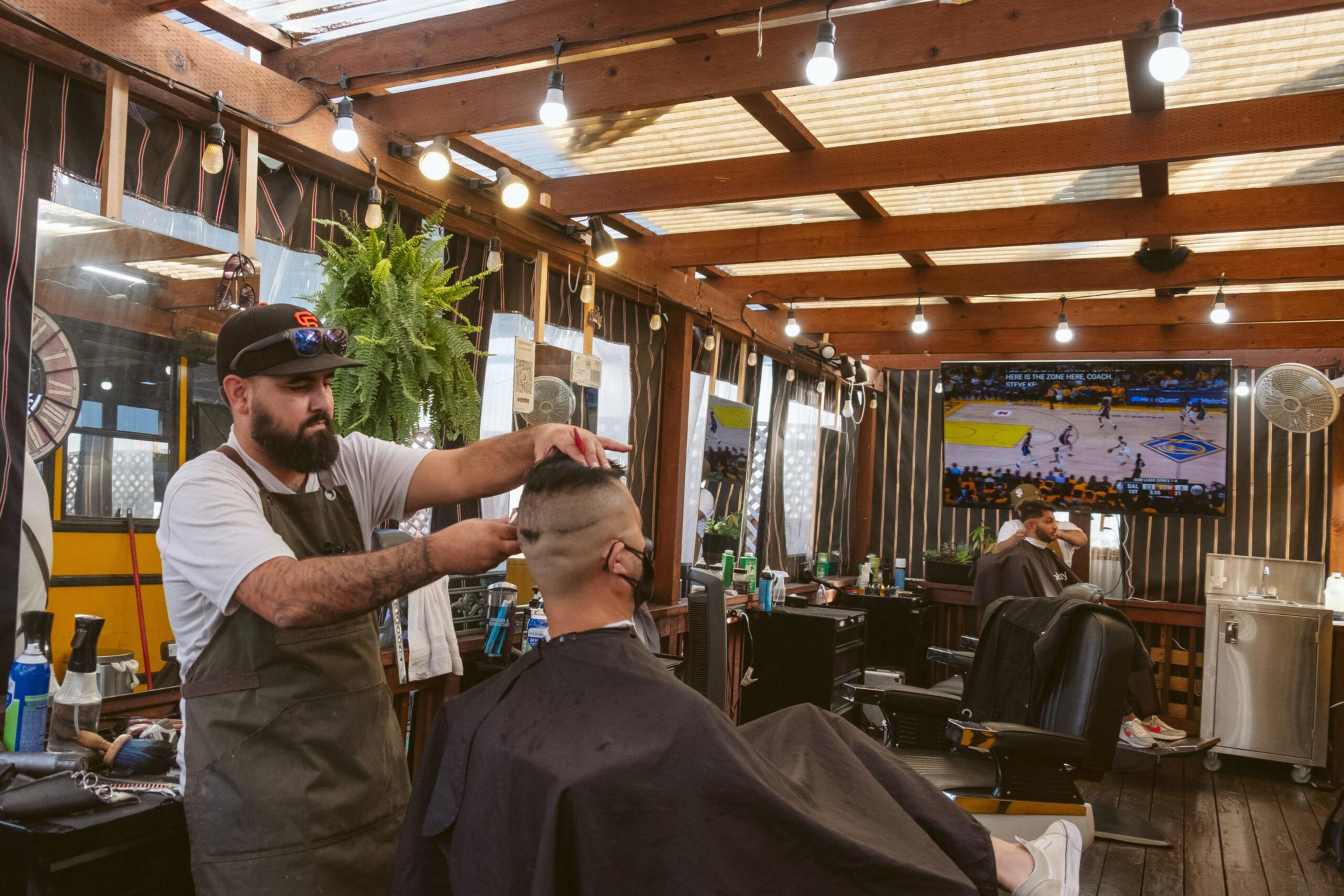 A barber wearing an apron and cap trims a customer's short hair inside a wooden, well-lit shop with hanging plants and a TV showing a basketball game.