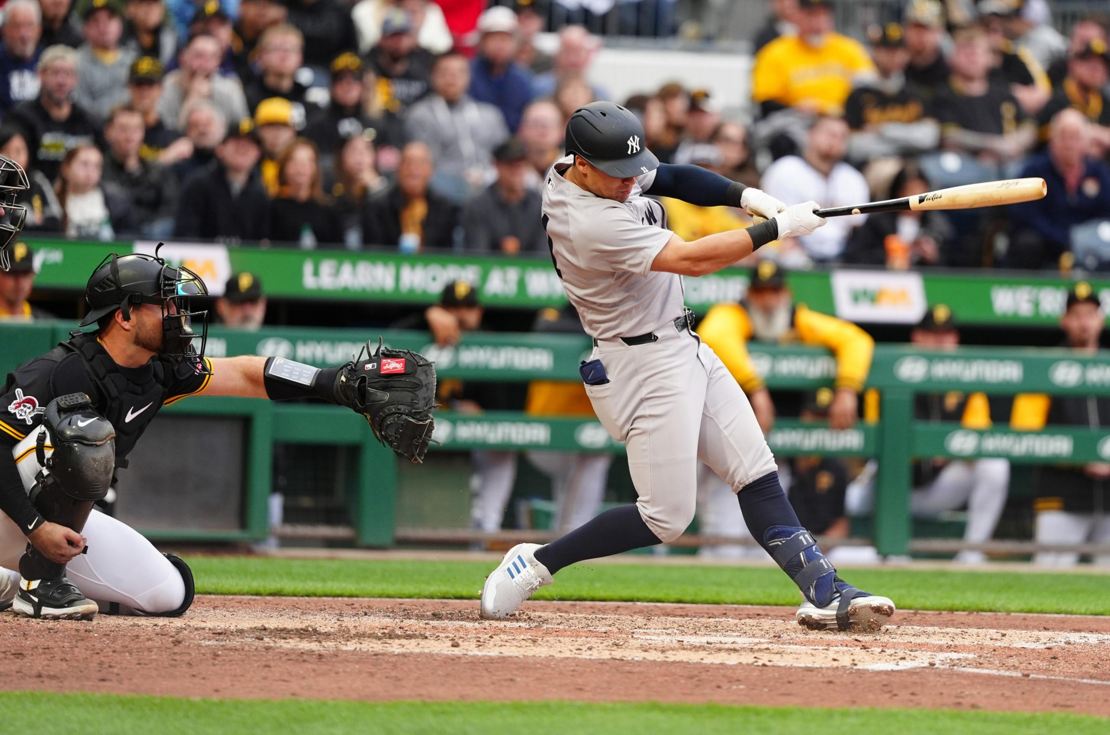 A baseball player in a gray uniform swings at a pitch while a black-clad catcher extends his glove; spectators in the background watch intently.